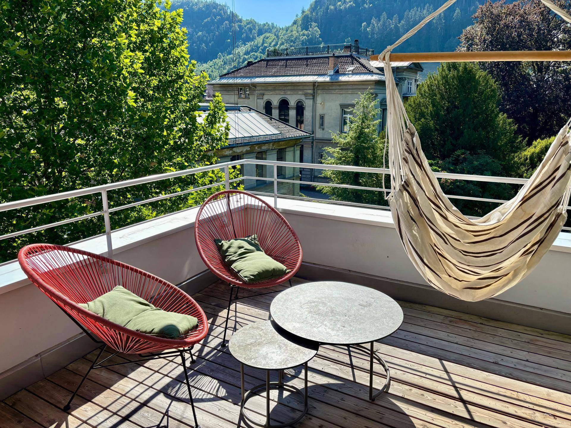 Geräumiger Balkon mit Holzfußboden, Hängematte und Sitzgelegenheiten, umgeben von viel Grün.