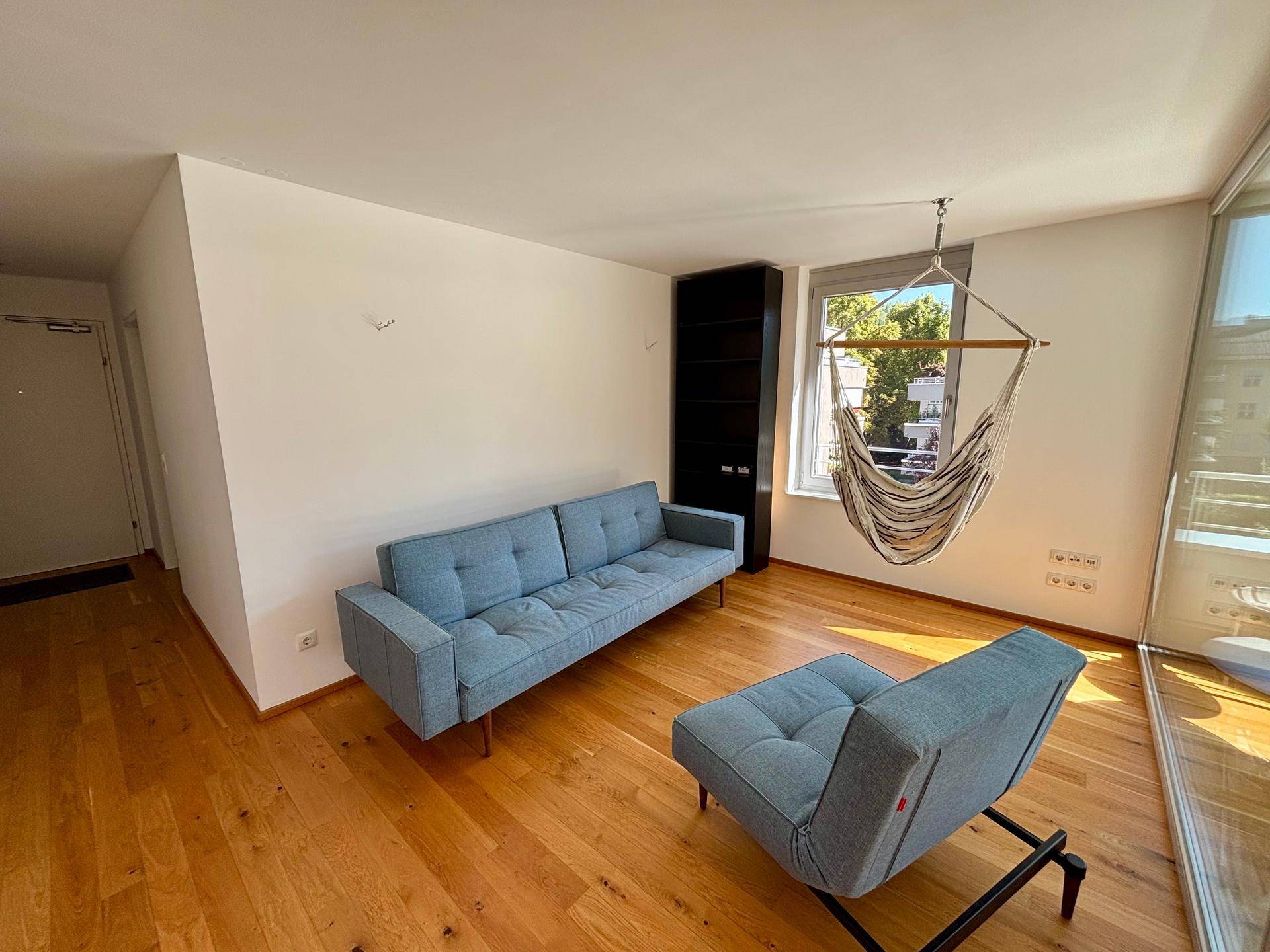 Helles Wohnzimmer mit Holzboden, modernem Sofa und einer Hängematte am Fenster.