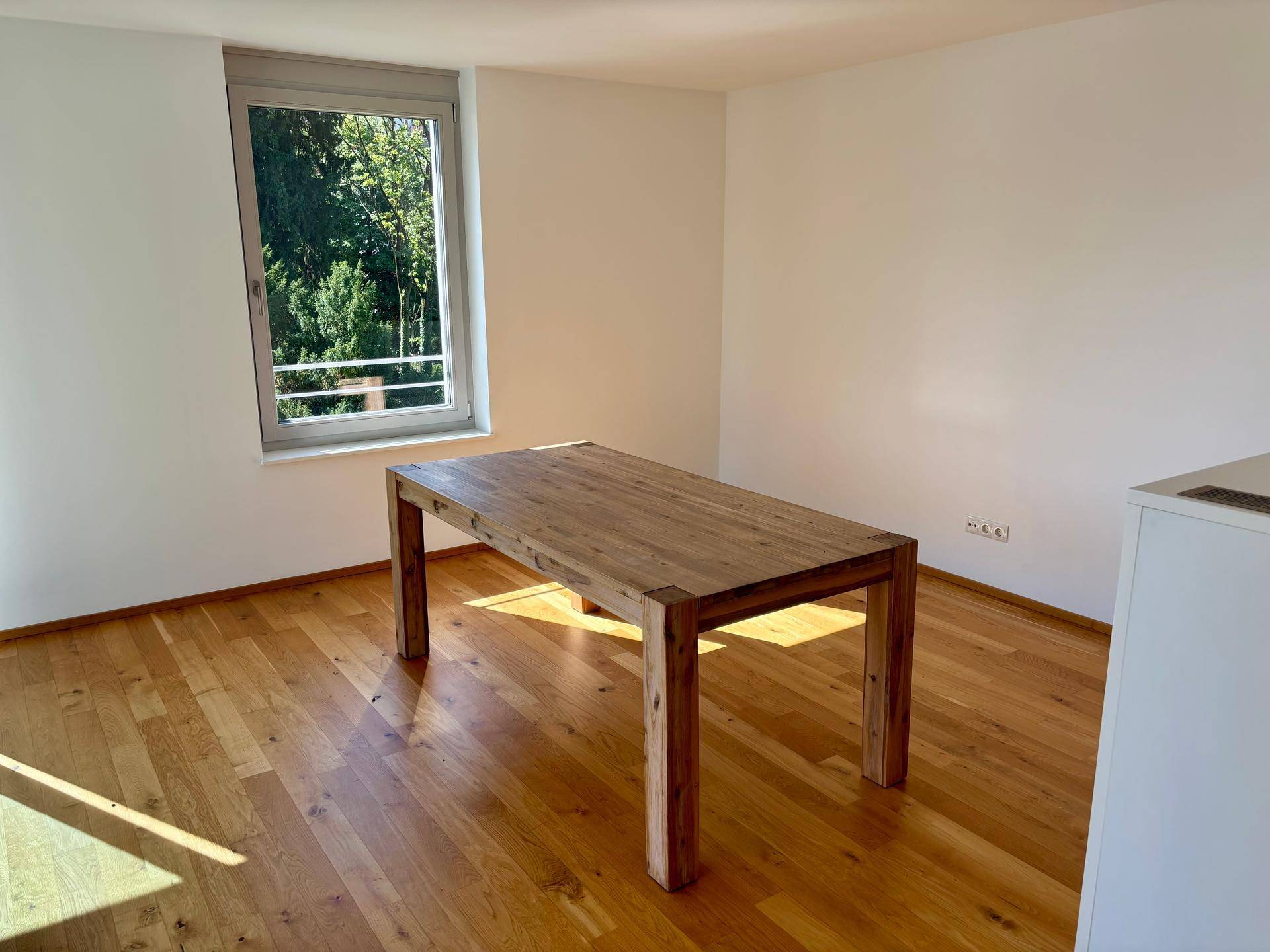 Essbereich mit großem Holztisch und Holzboden, hell durch das Fenster mit Blick ins Grüne.