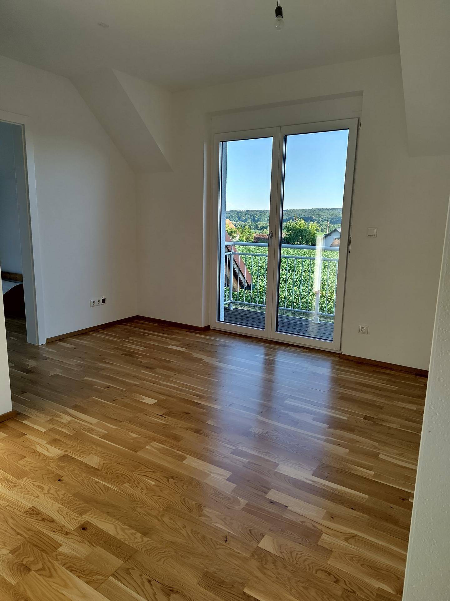 Helles Schlafzimmer mit Holzboden, Dachschräge und Zugang zu einem Balkon mit weitem Blick ins Grüne.