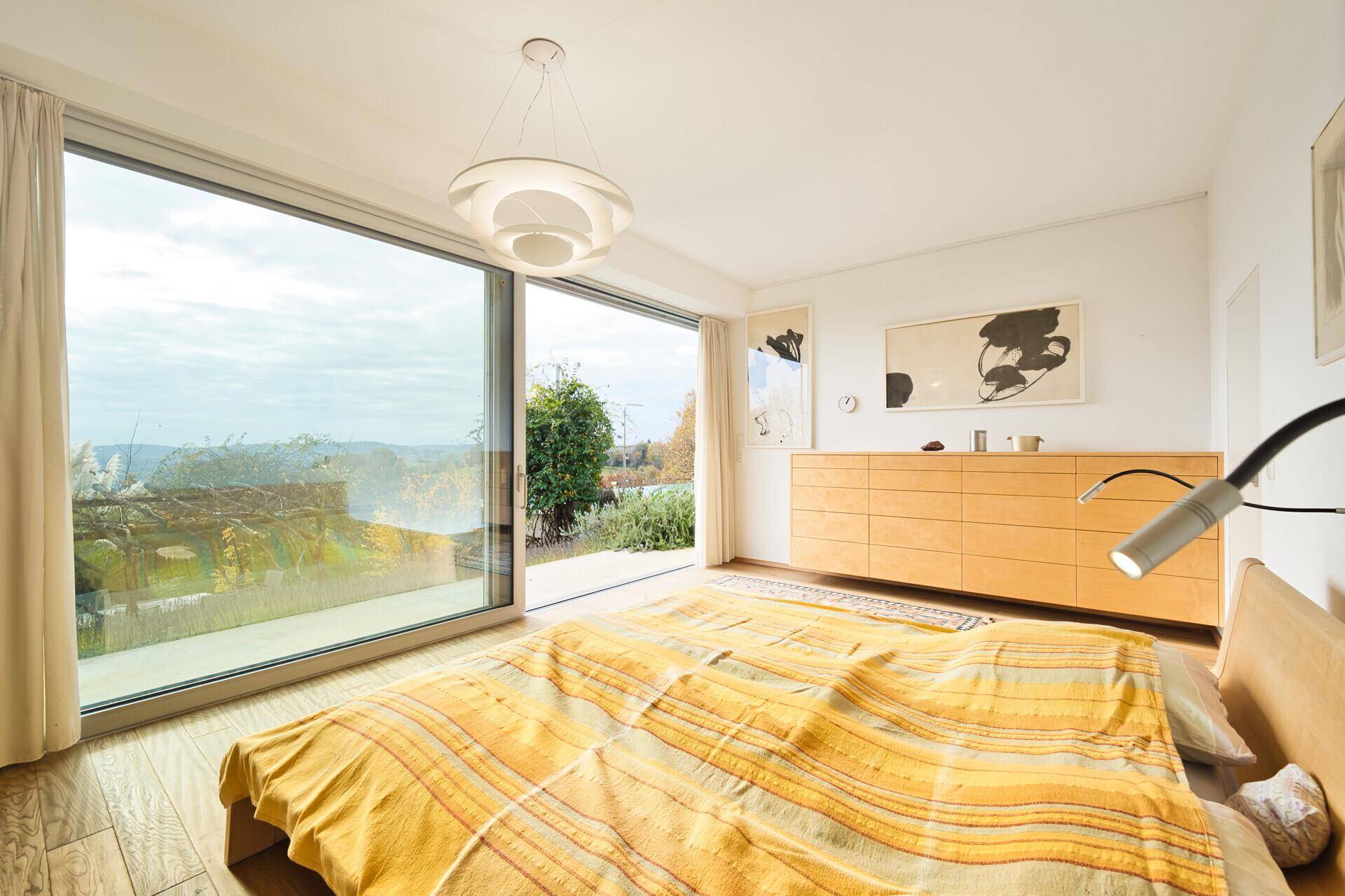 Helles Schlafzimmer mit Holzboden, großen Fenstern und direktem Zugang zum Garten mit grüner Aussicht.