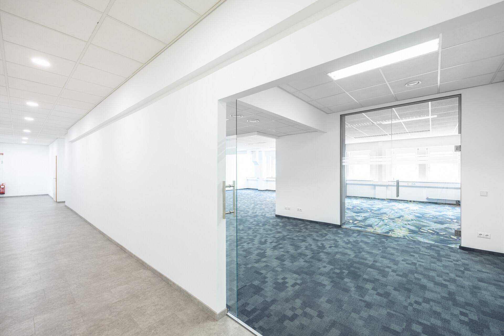 Langer Bürogang mit grauen Fliesenböden und Zugang zu einem Teppichboden-Büro durch eine Glastür.