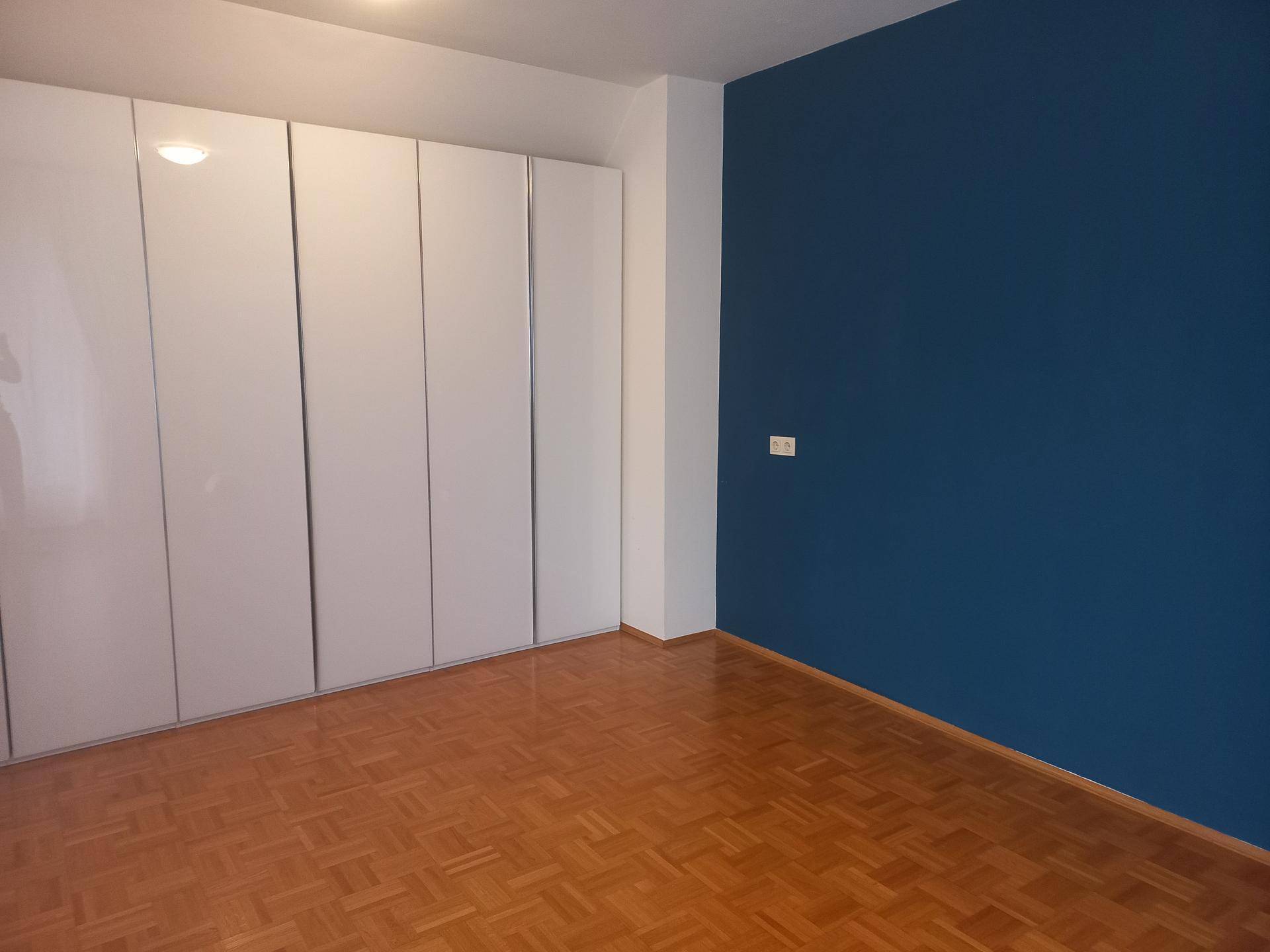 Schlafzimmer mit großem weißen Einbauschrank, blauer Akzentwand und Parkettboden.
