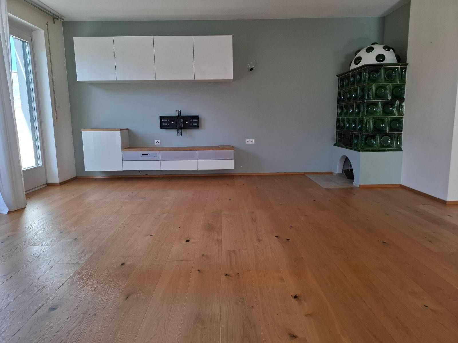 Geräumiges Wohnzimmer mit Holzboden, modernem Wandregal und traditionellem Kachelofen.