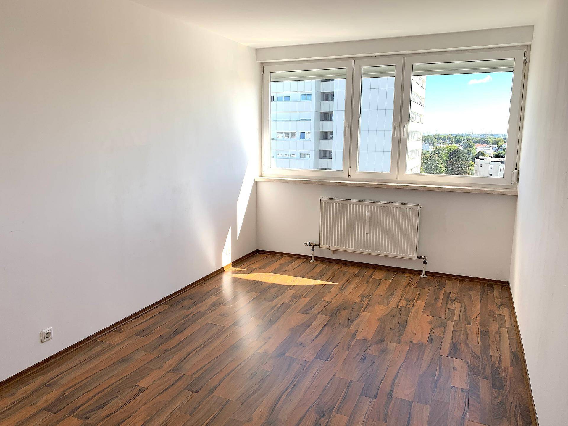 Helles, leeres Zimmer mit Laminatboden und großem Fenster mit Stadtblick.