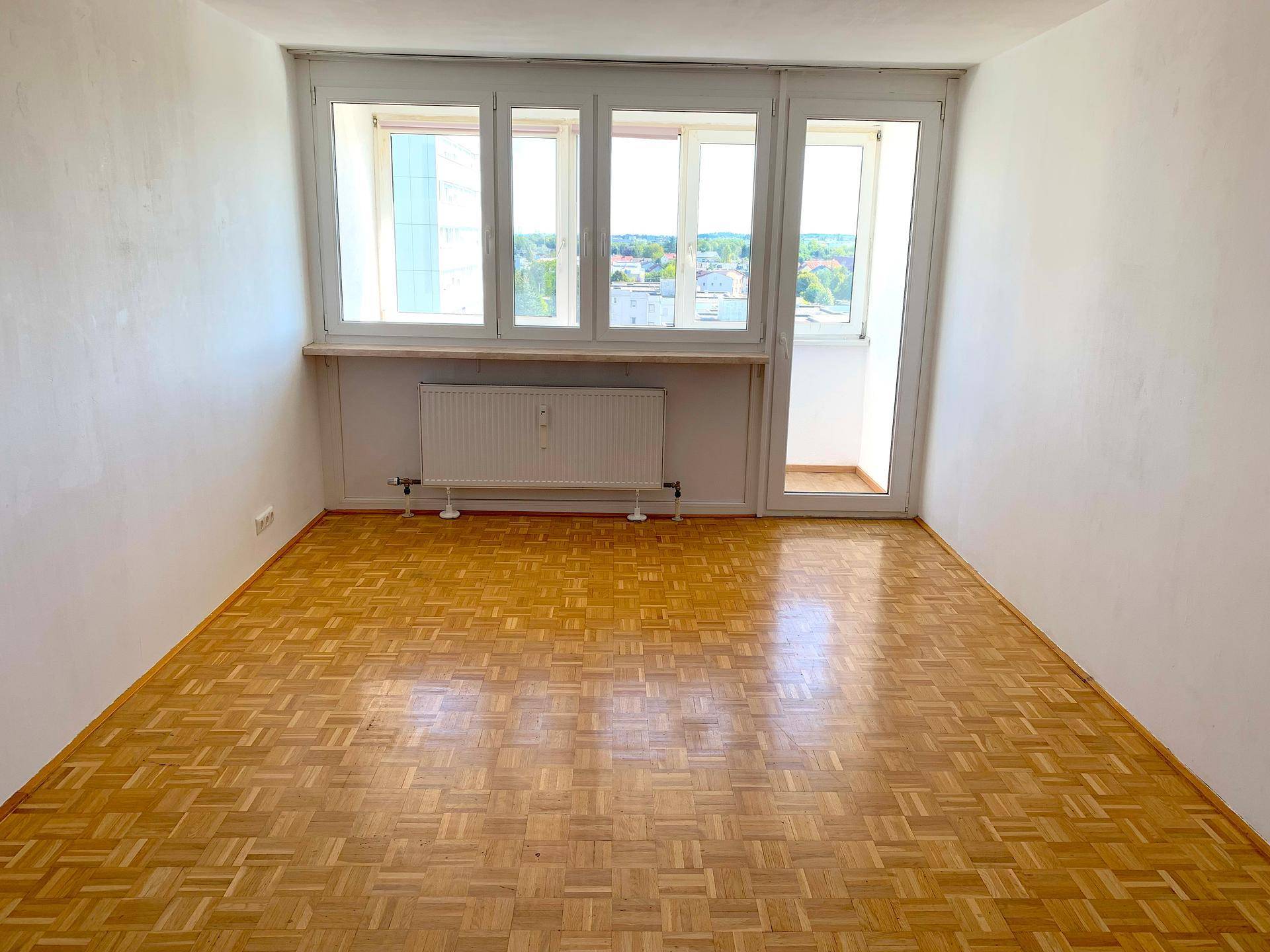 Helles, leeres Zimmer mit Fischgrätparkett, großem Fenster und Zugang zu einem kleinen Balkon.