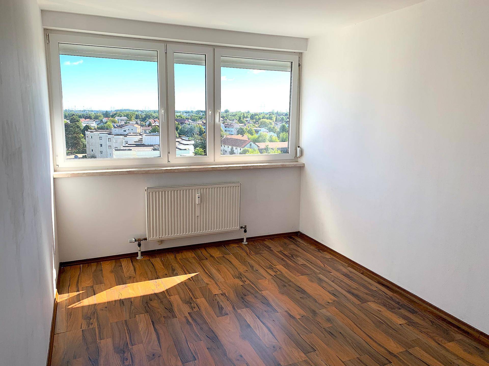 Leeres Zimmer mit Laminatboden und Fenster, das einen weiten Blick über die Umgebung bietet.
