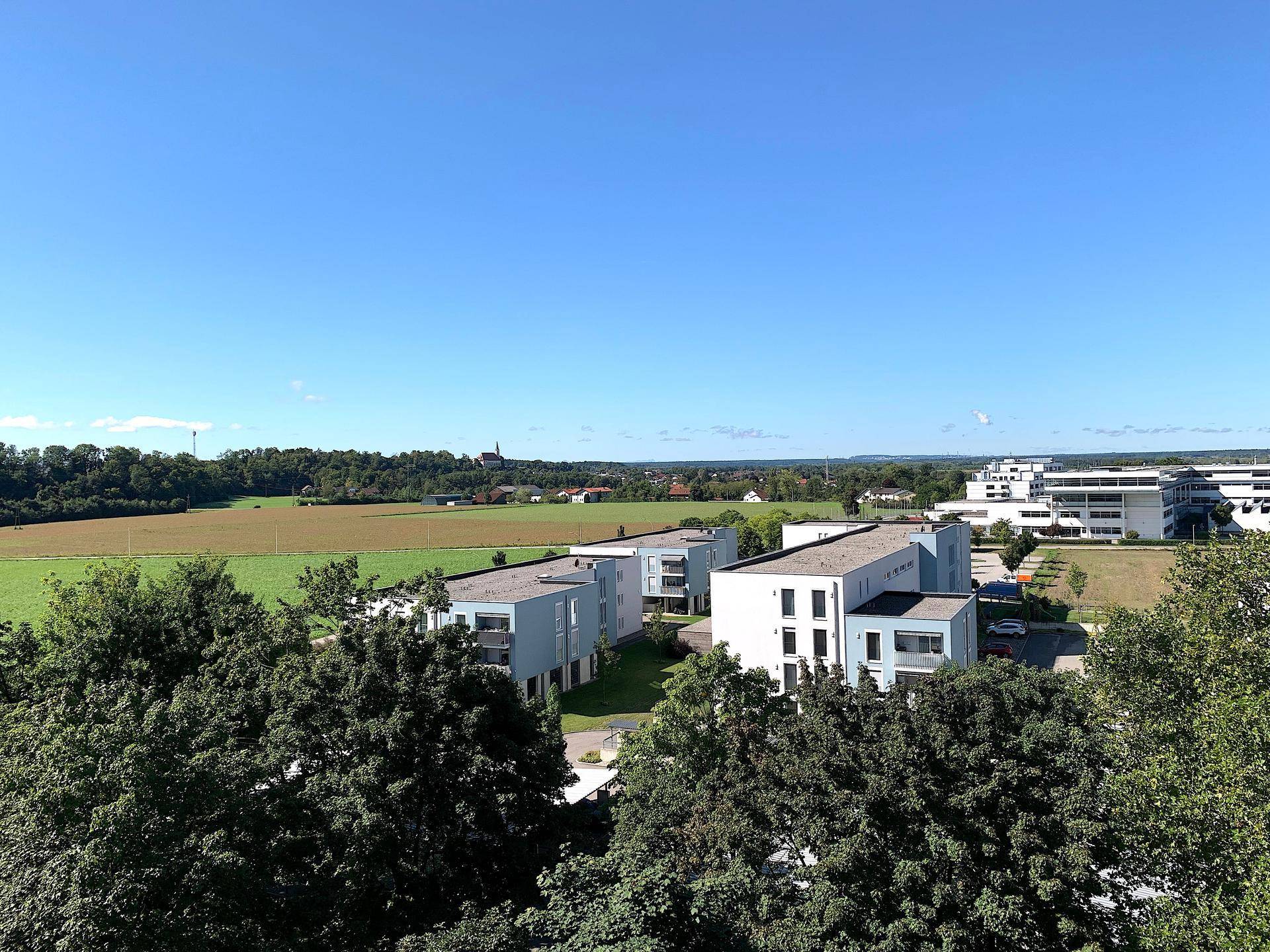 Panoramablick auf moderne Wohngebäude, weite Felder und eine grüne Hügellandschaft unter klarem Himmel.