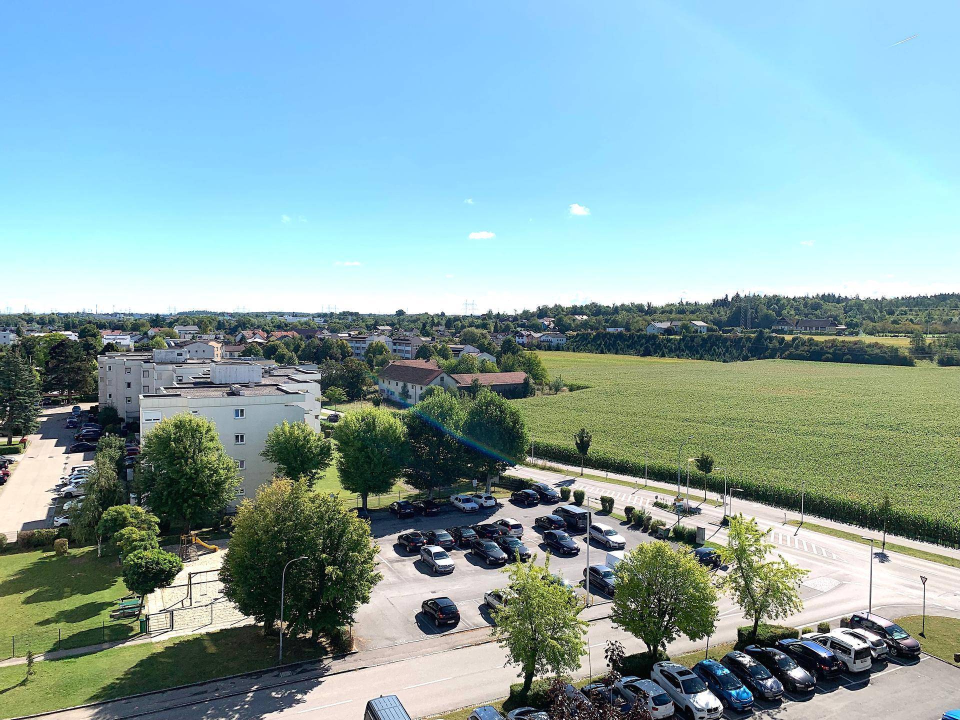 Weitläufiger Blick über einen Parkplatz, grüne Felder und eine Wohnsiedlung in der Ferne.