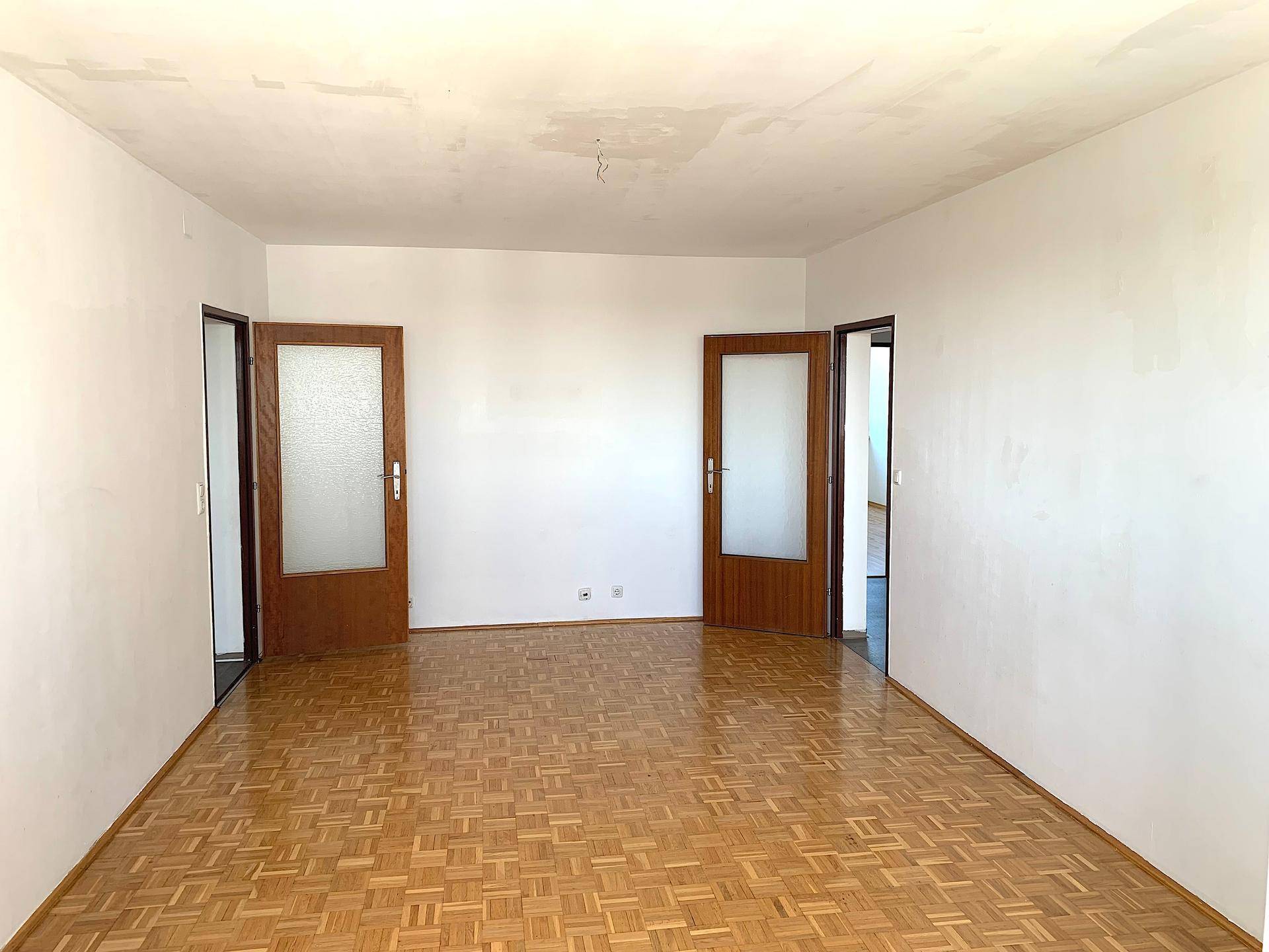 Geräumiges, leeres Zimmer mit Fischgrätparkett und zwei Holztüren.