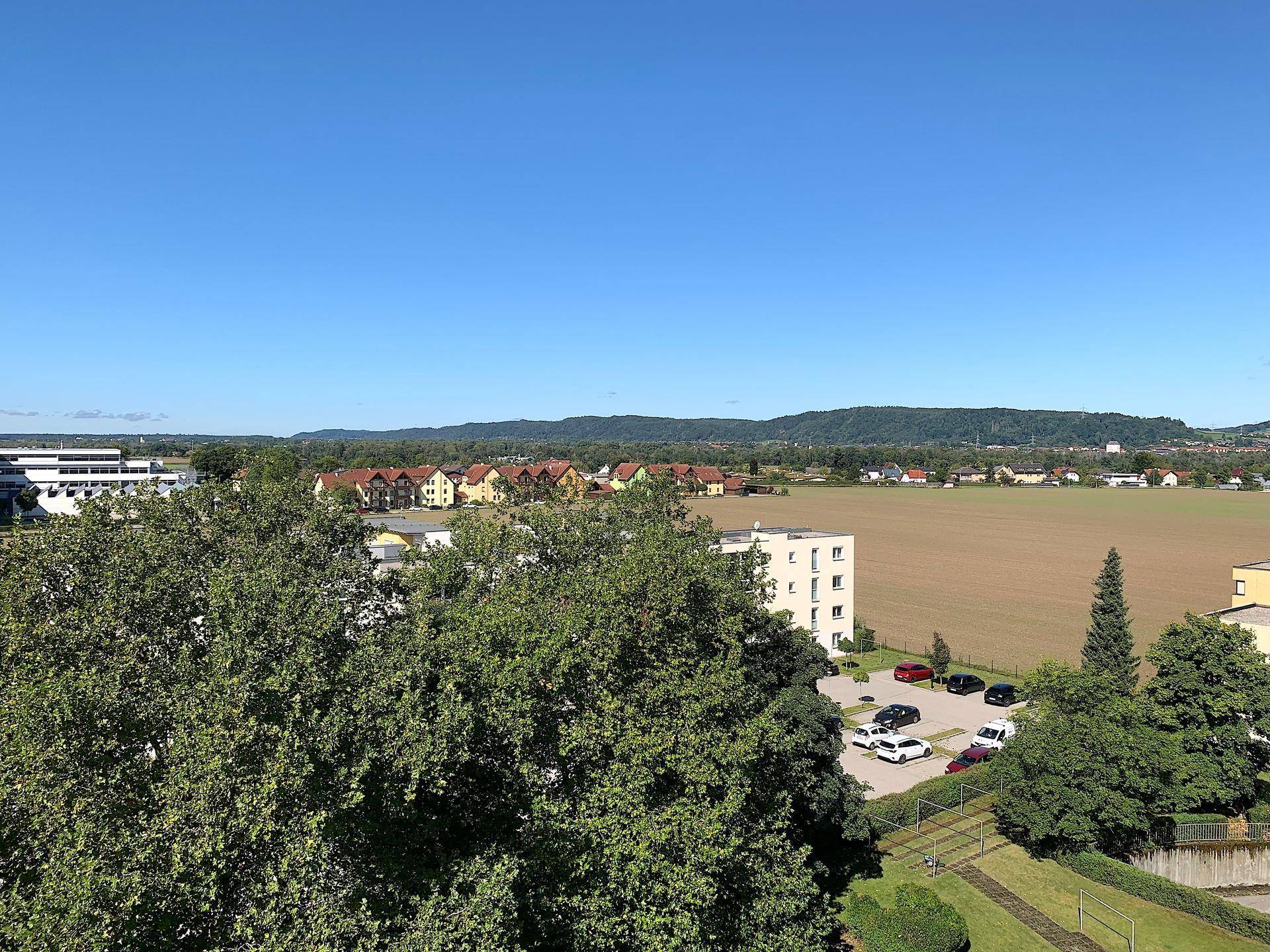 Weitläufiger Blick über grüne Felder, Wohnsiedlungen und eine hügelige Landschaft am Horizont.