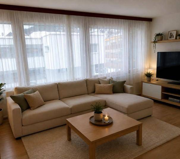 Helles Wohnzimmer mit beigem Ecksofa, Couchtisch und großen Fenstern mit transparenten Vorhängen.