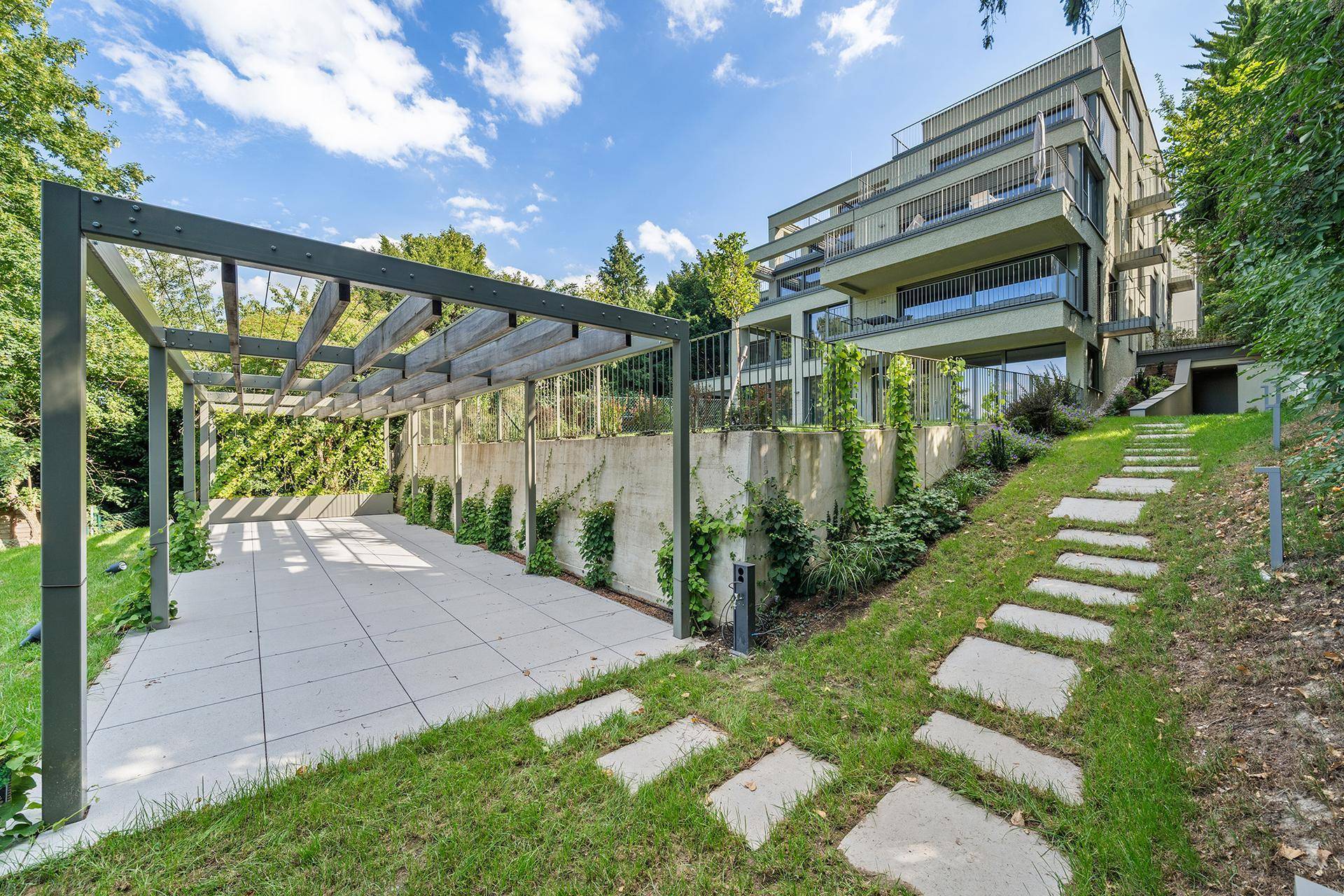 Moderner Gebäudekomplex mit Terrassen und einem Gartenbereich, inklusive Pergola und Steinstufen.