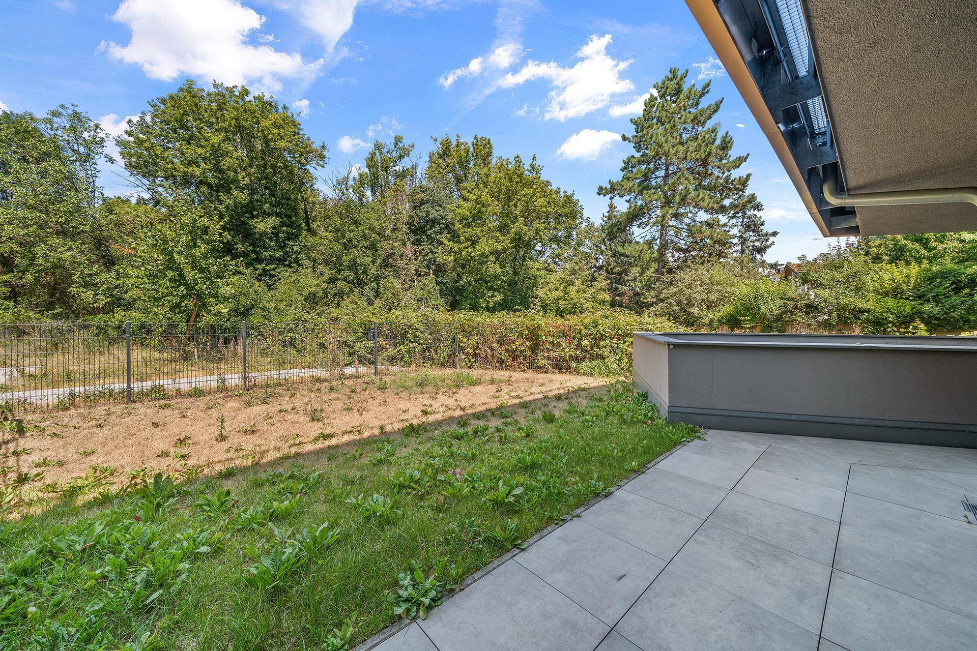 Sonniger Terrassenbereich mit Fliesenboden und angrenzendem Garten, umgeben von viel Grün.