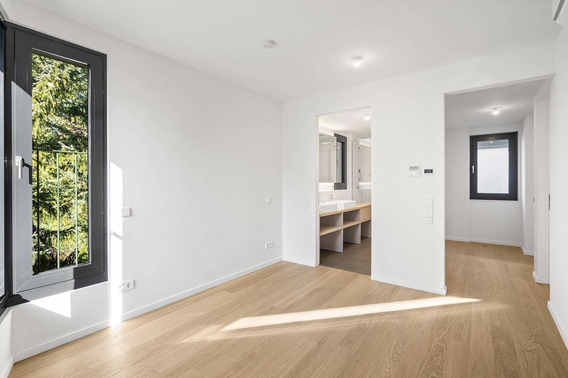 Helles Schlafzimmer mit Holzboden, großem Fenster und Zugang zum modernen Badezimmer.