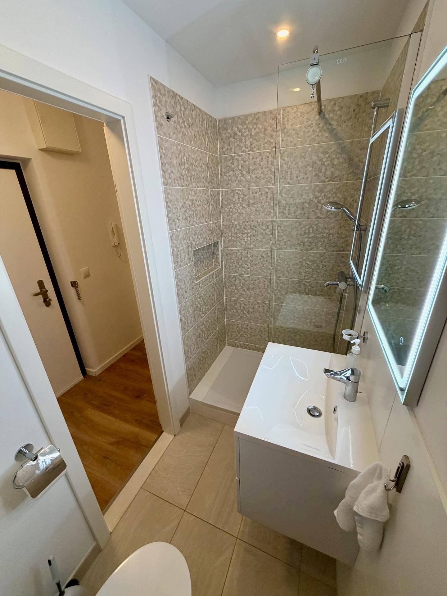 Modernes Badezimmer mit Duschkabine, Waschtisch und beleuchtetem Spiegel, Blick zum Flur.
