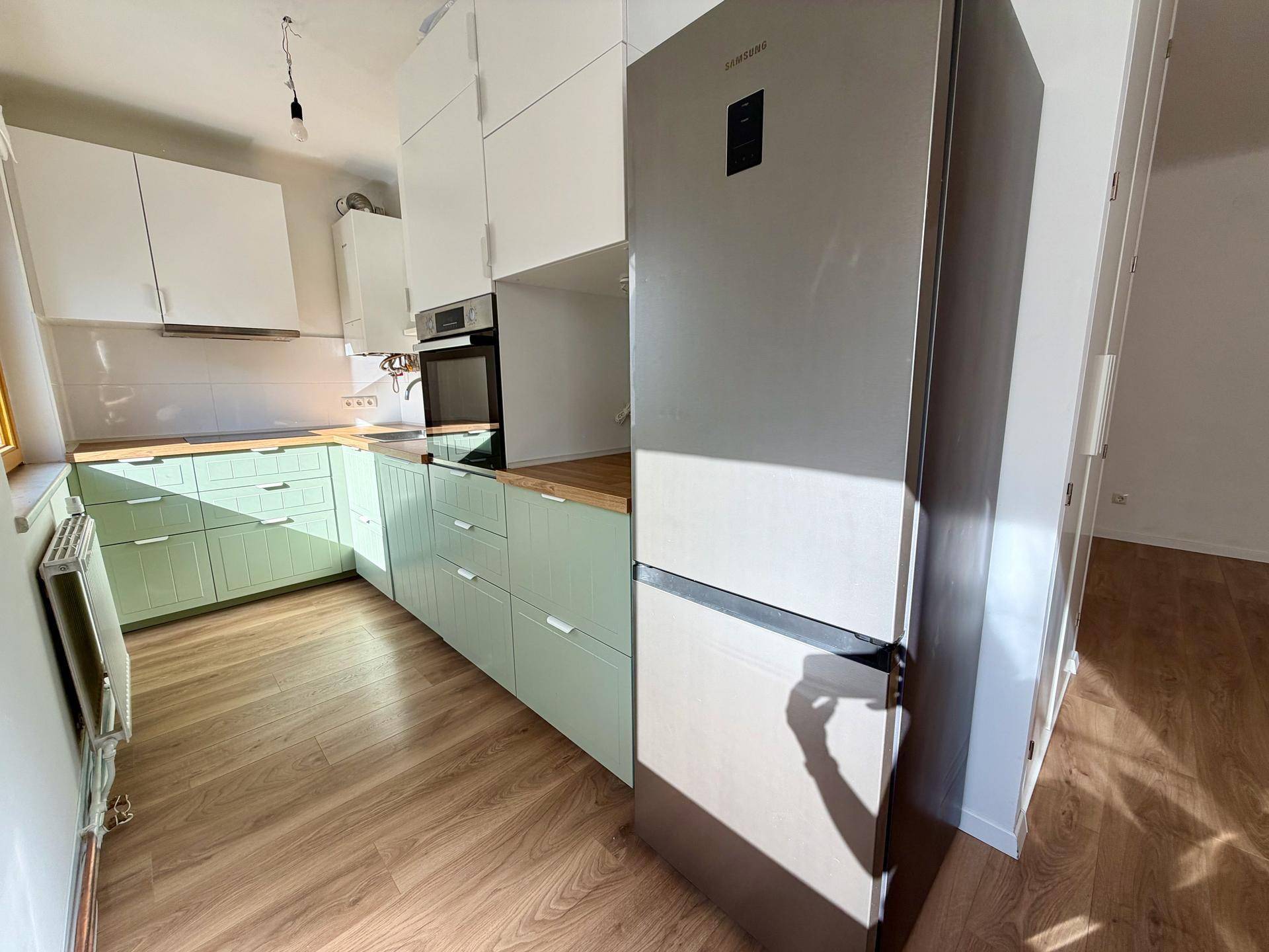 Gesamtaufnahme der modernen Küche mit Kühlschrank, Backofen und hellgrünen Schränken.