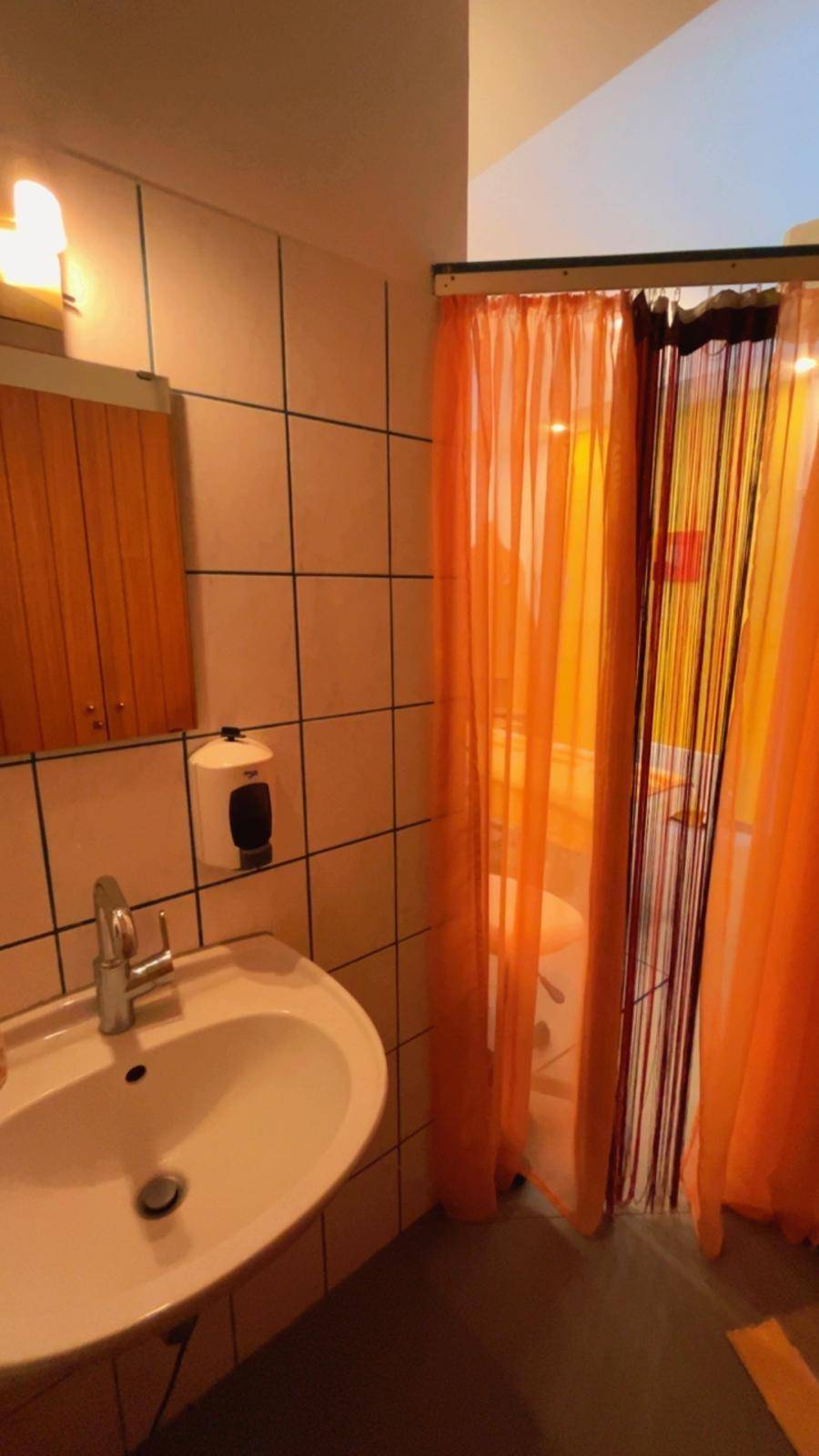 Kleines Badezimmer mit Waschbecken, Spiegel und einer Duschkabine hinter einem orangefarbenen Vorhang.