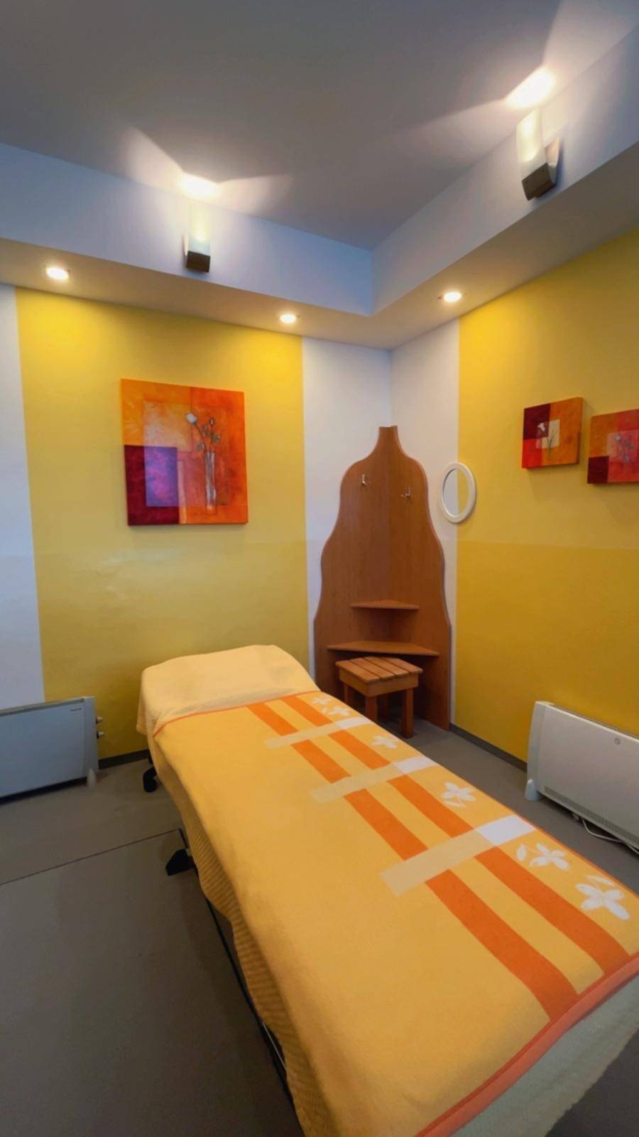 Heller Behandlungsraum mit gelben Wänden, moderner Kunst, einer Massageliege und indirekter Beleuchtung.