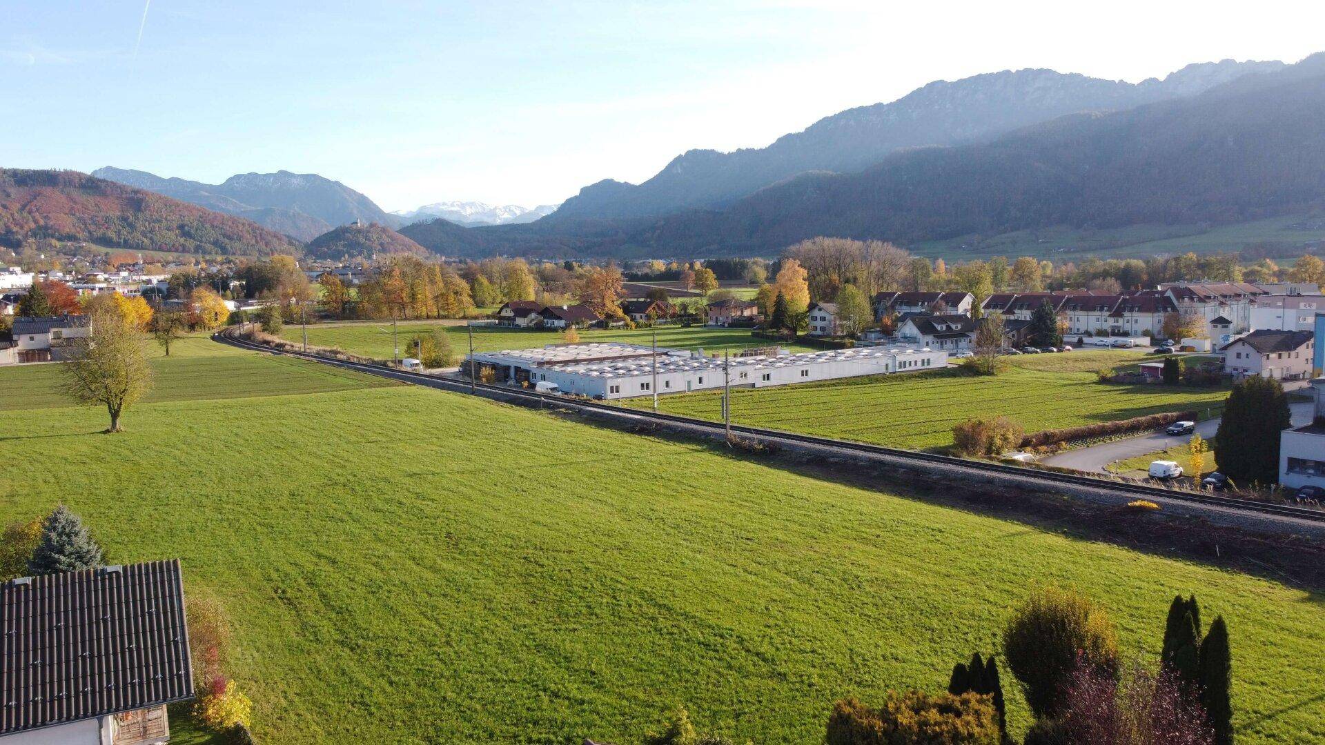 Luftaufnahme zeigt grüne Felder, eine Bahntrasse und eine Siedlung vor einer beeindruckenden Bergkette.