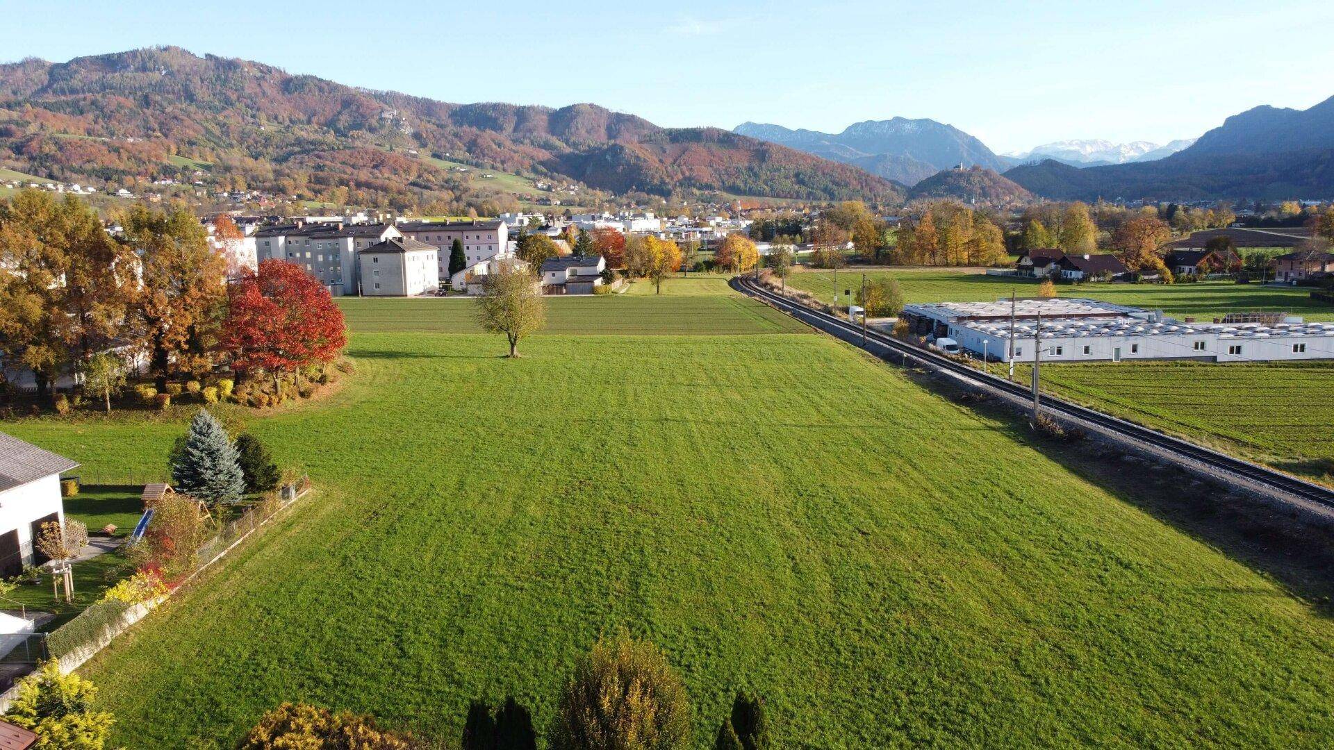 Panoramablick aus der Vogelperspektive auf weite grüne Wiesen, eine Bahnlinie und eine Stadt mit herbstlichen Bäumen.