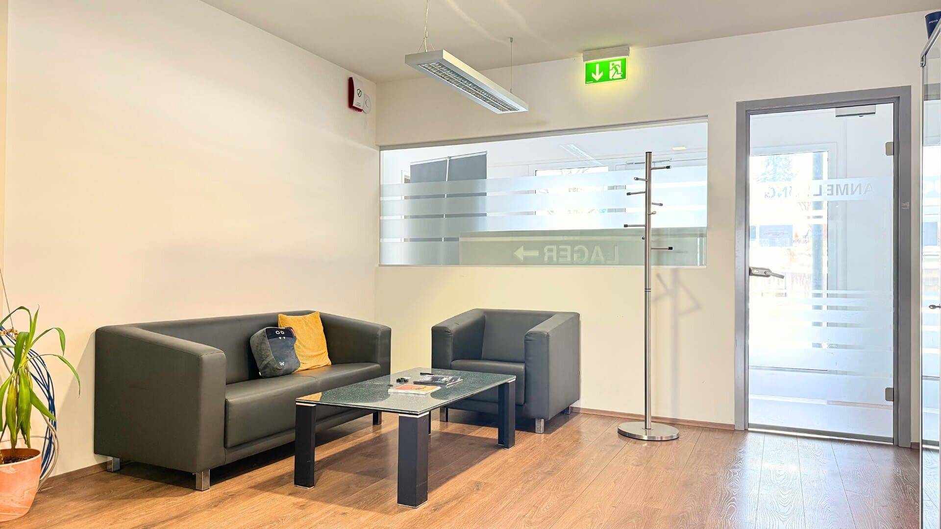Gemütlicher Wartebereich mit Sofa, Sessel und Couchtisch, ergänzt durch moderne Glastüren mit Sichtschutz.