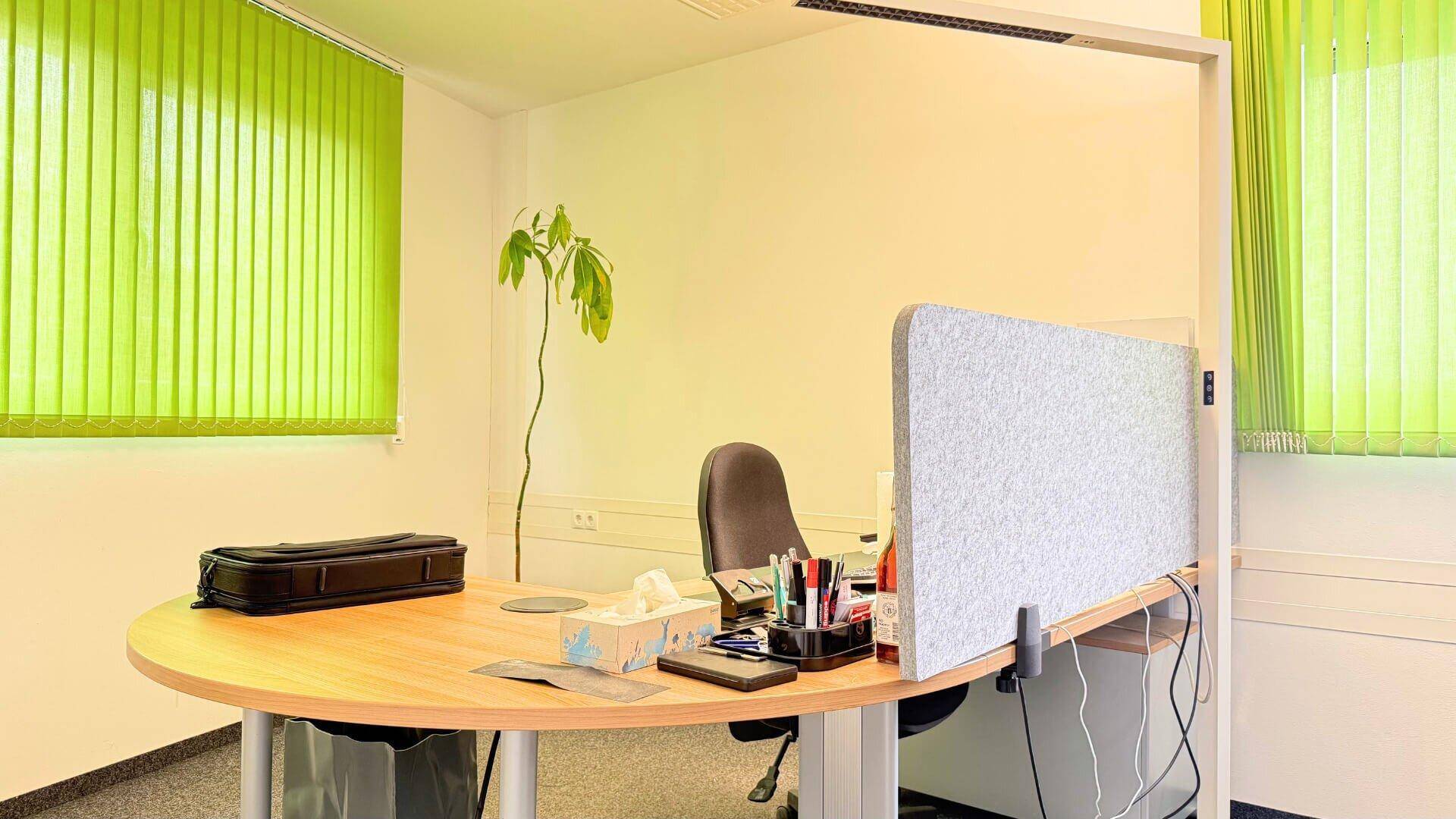 Einzelner Arbeitsplatz in einem Büro mit Schreibtisch, Bürostuhl und hellgrünen Jalousien an den Fenstern.