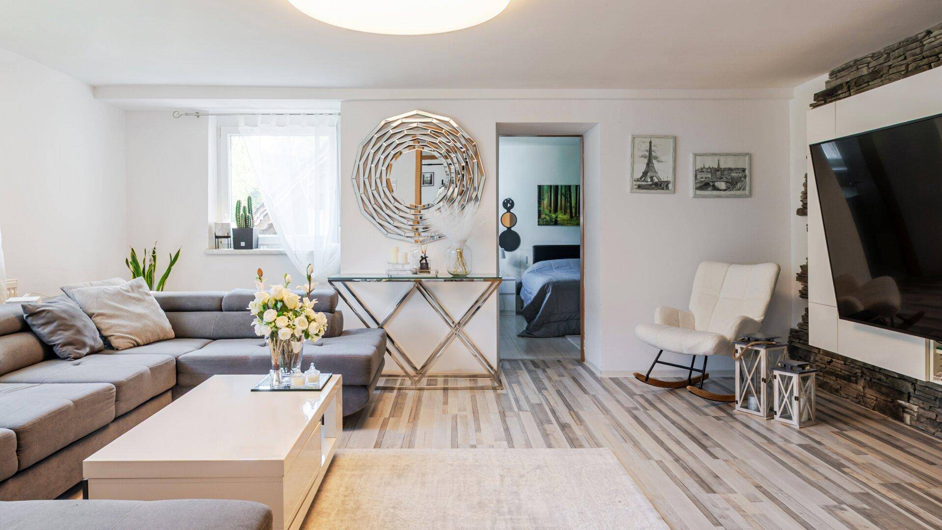 Helles Wohnzimmer mit modernem Sofa, stilvollem Spiegel und Zugang zu einem weiteren Raum.