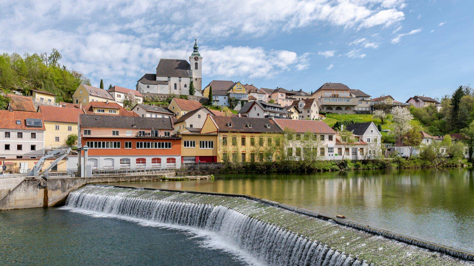 Malerische Stadtansicht mit einer Kirche auf dem Hügel, einem Fluss und einem Wehr im Vordergrund.
