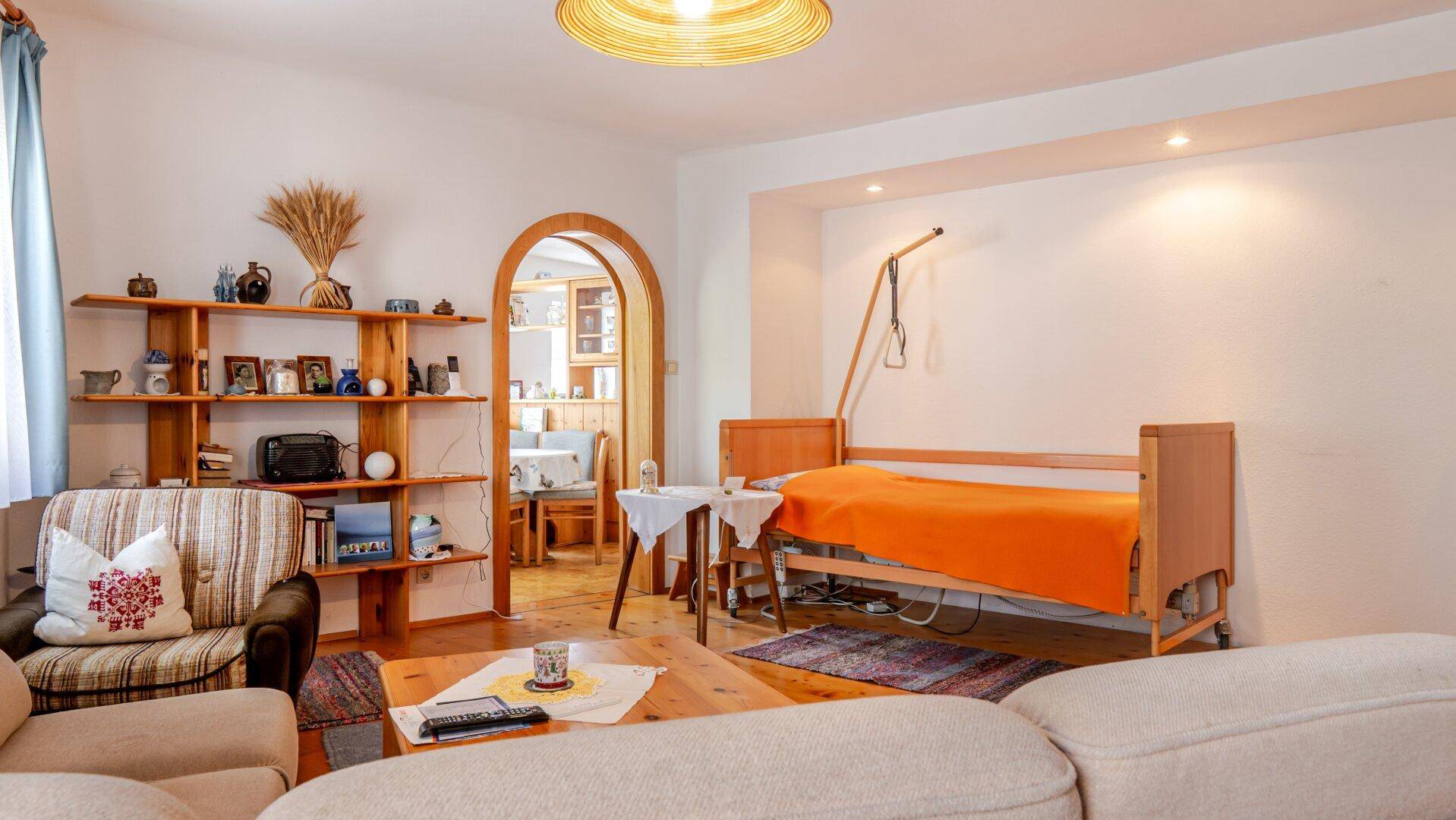 Wohnzimmer mit Pflegebett, Holzregal und Durchgang zu einem weiteren Raum.