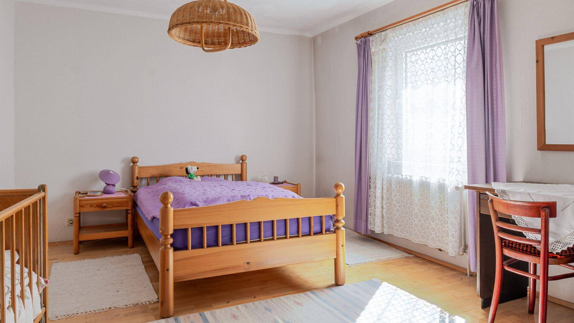 Helles Schlafzimmer mit Holzbett, lila Bettwäsche und Kinderbett.