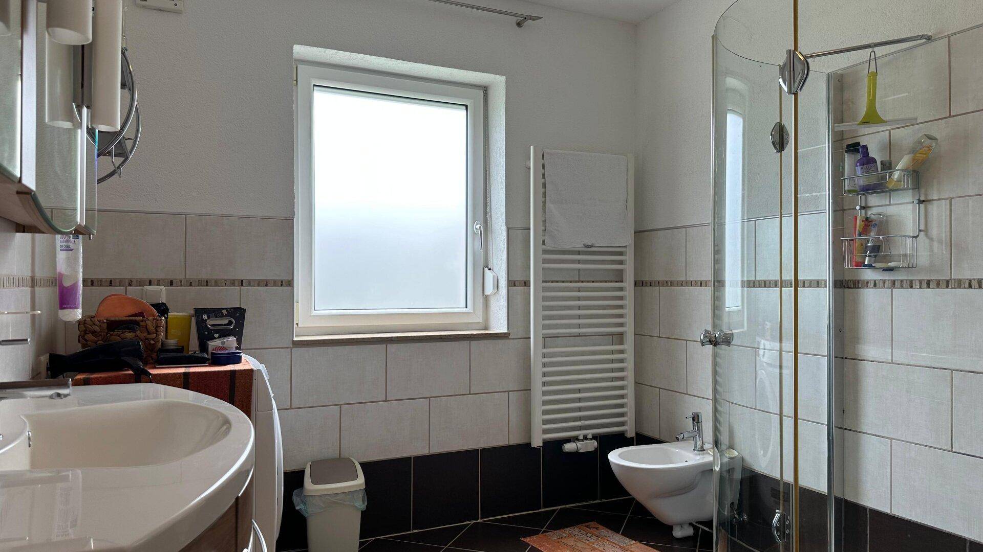 Badezimmer mit Duschkabine, Bidet, Waschbecken und Fenster, ausgestattet mit Handtuchheizkörper.