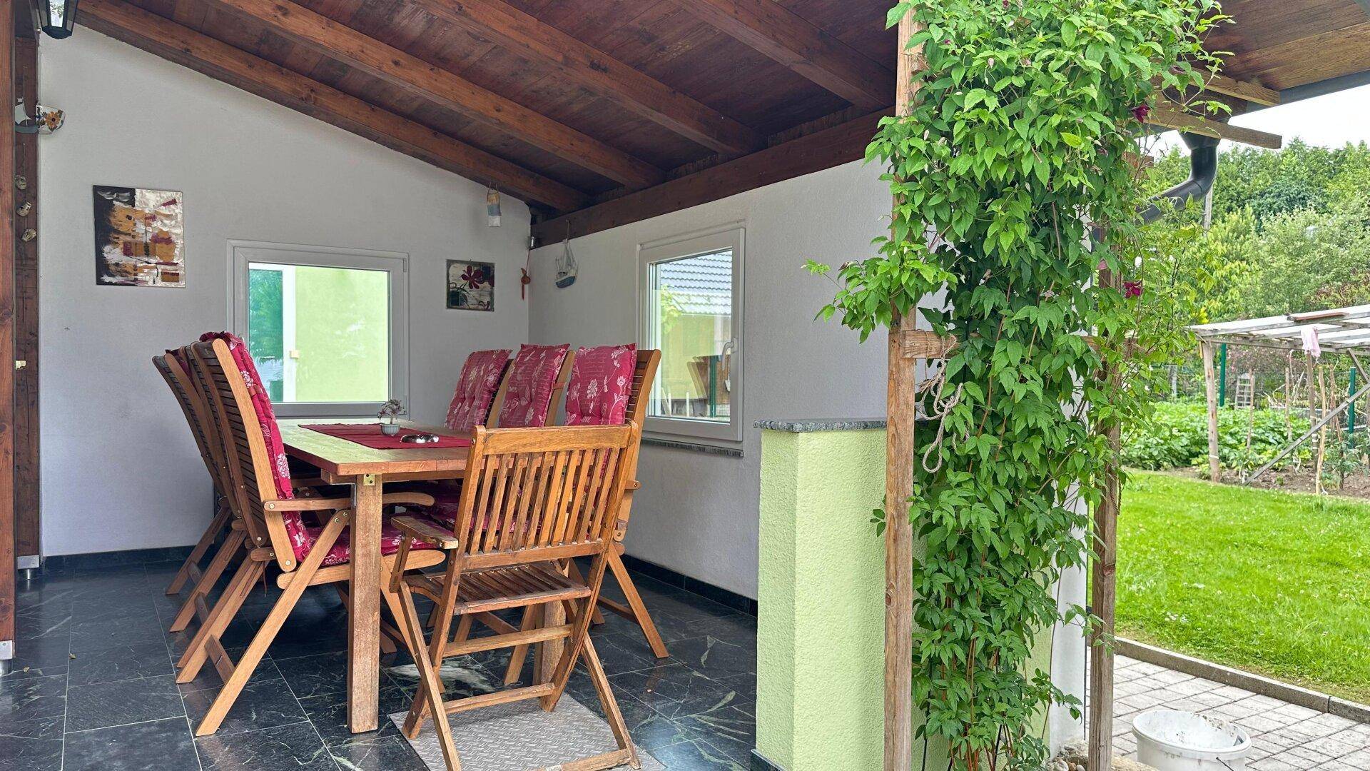 Überdachter Essbereich im Freien mit Holztisch und Stühlen, mit Blick auf den Garten.