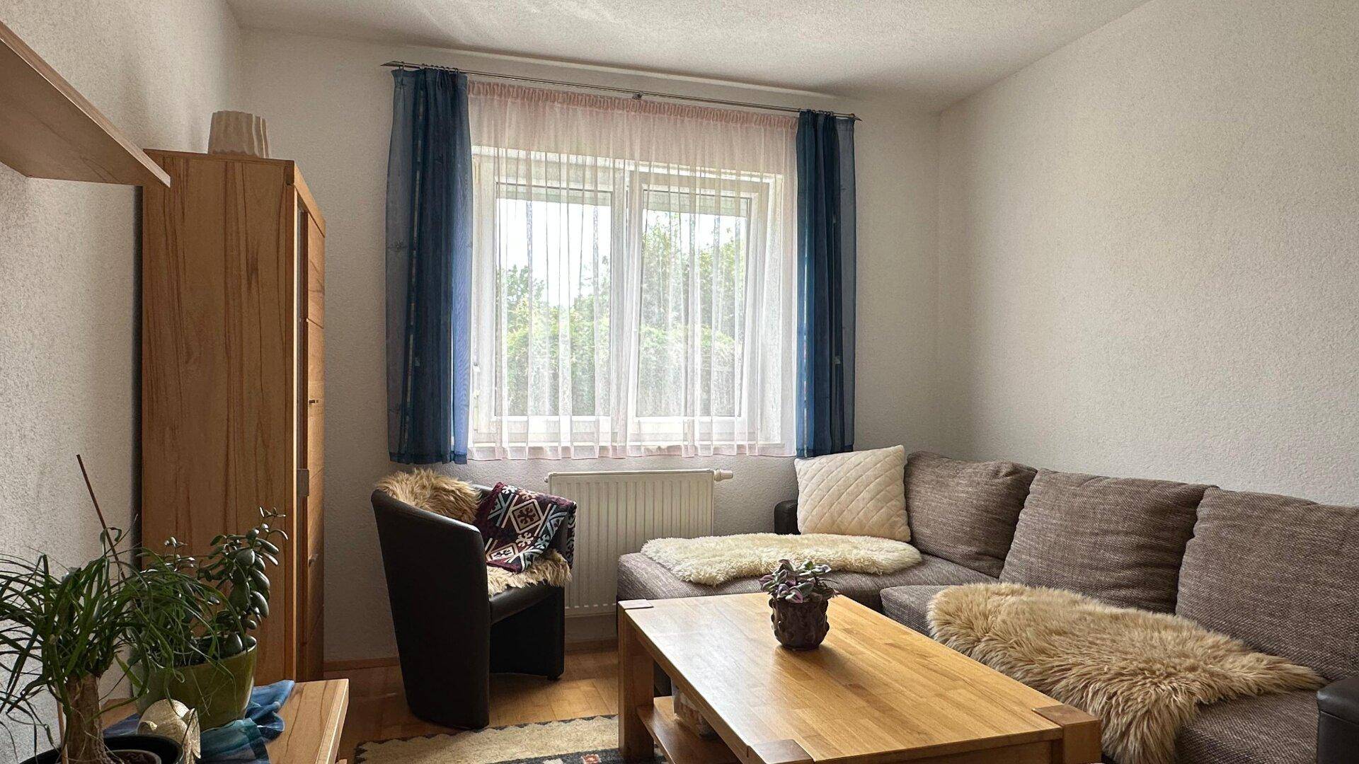 Kleiner Wohnbereich mit braunem Sofa, Holztisch und Fenster mit blauen Vorhängen.
