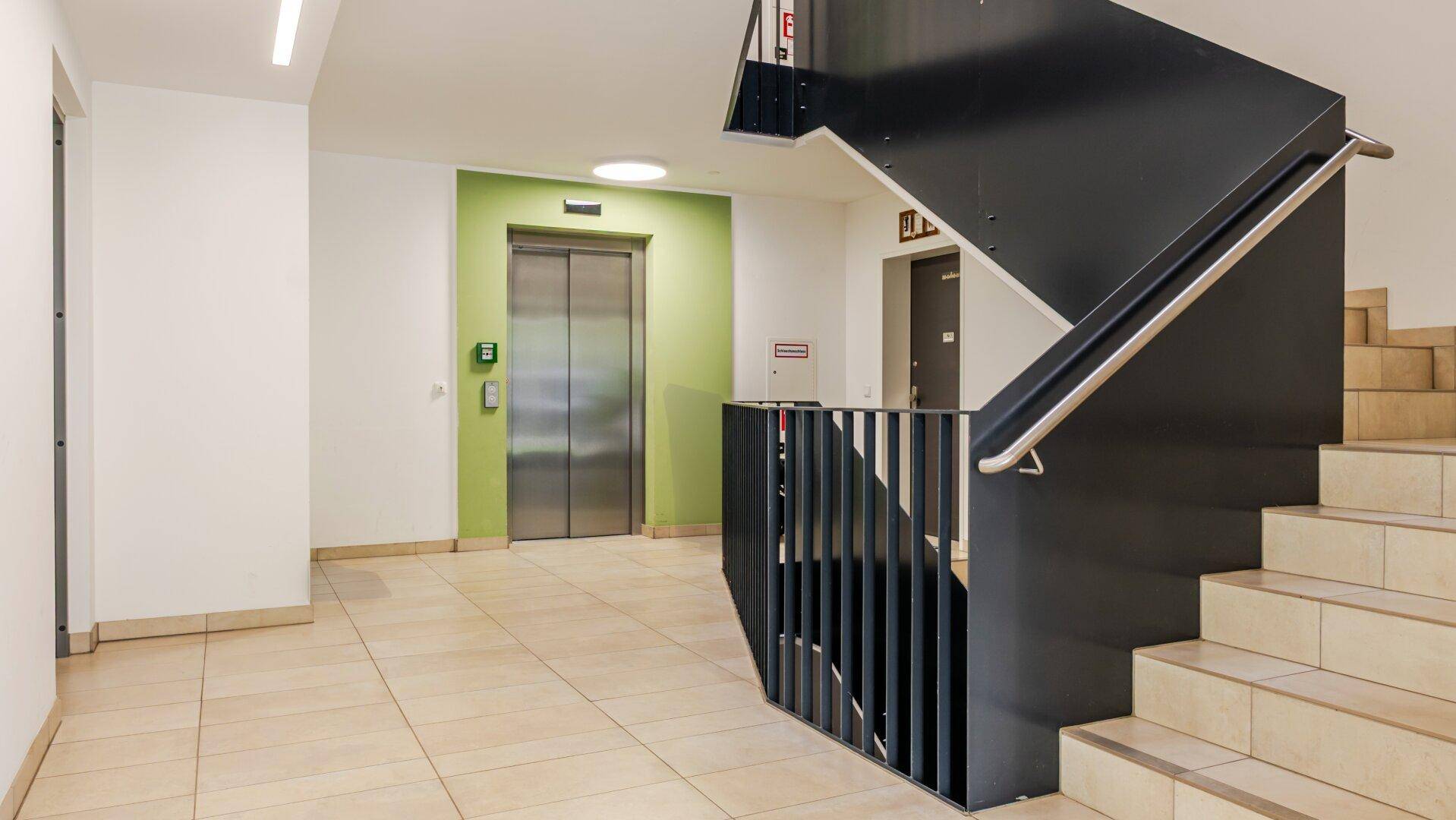 Moderner Treppenaufgang mit Aufzug und hellem Fliesenboden in einem Mehrfamilienhaus.