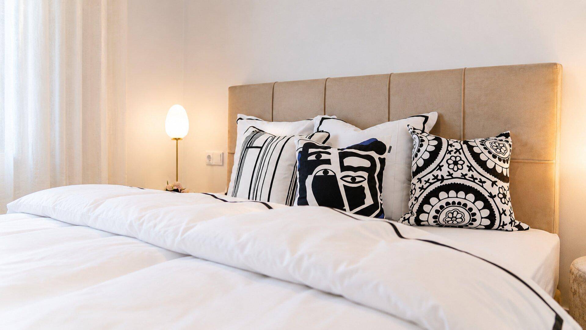Gemütliches Schlafzimmer mit beigem Kopfteil und dekorativen Kissen in Schwarz-Weiß-Mustern.