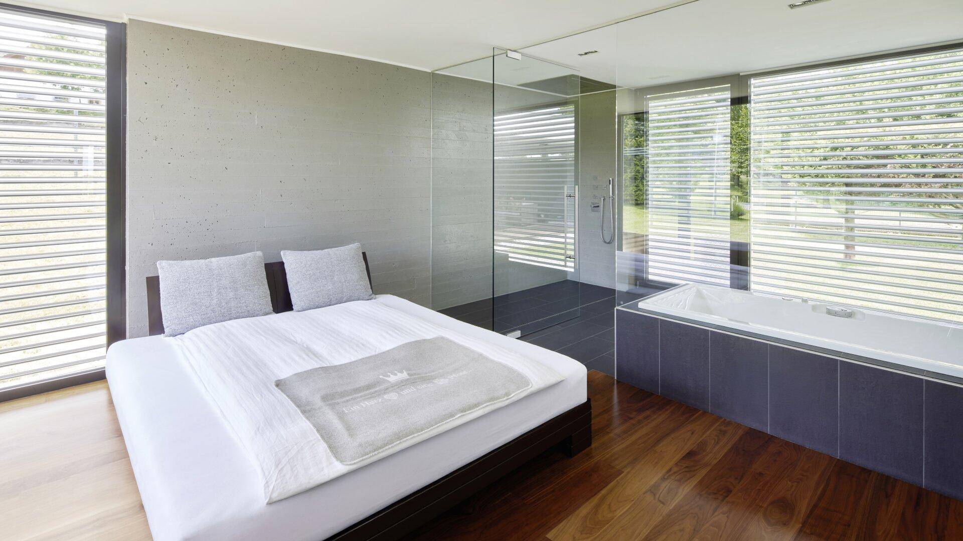 Schlafzimmer mit Holzboden, integrierter Dusche und Badewanne sowie großen Fenstern mit Jalousien.