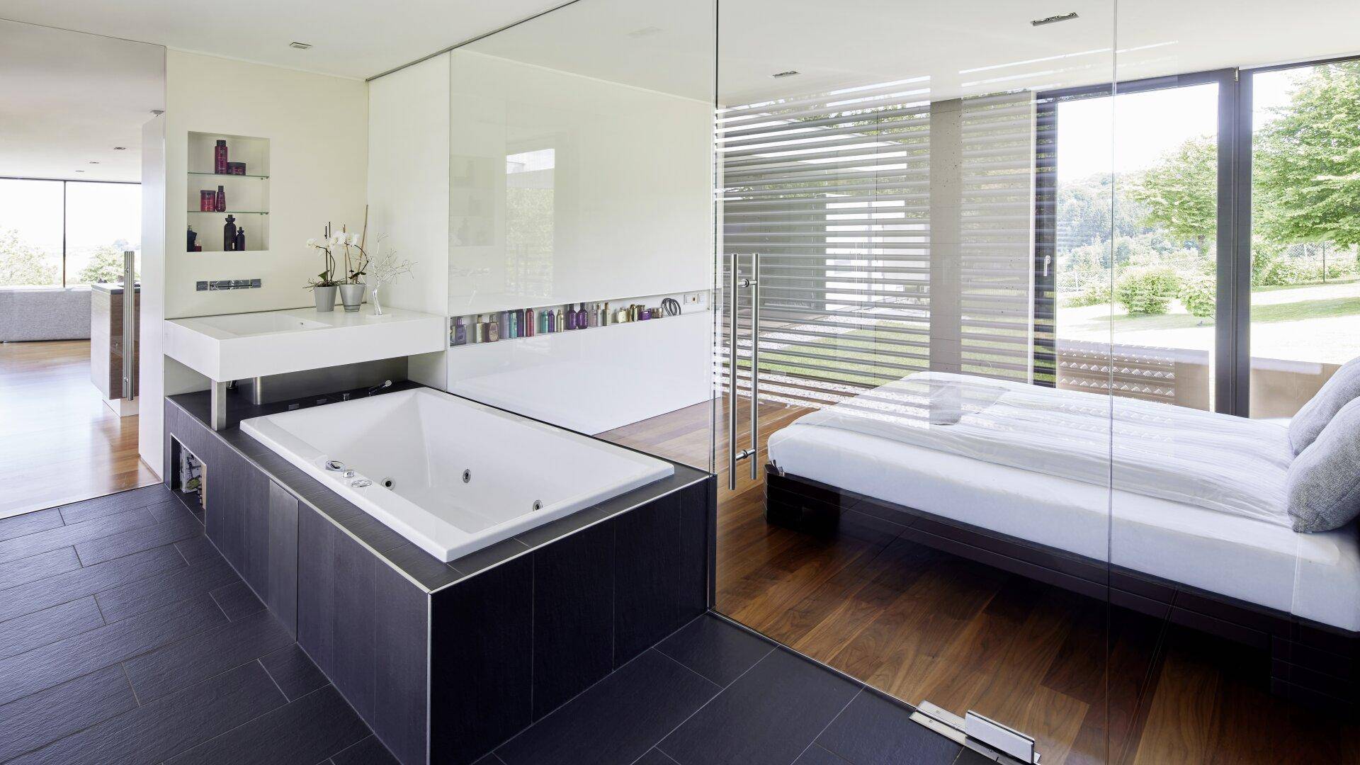 Offenes Schlafzimmer mit Badewanne, Doppelwaschbecken und Blick ins Grüne durch Glasabtrennung.