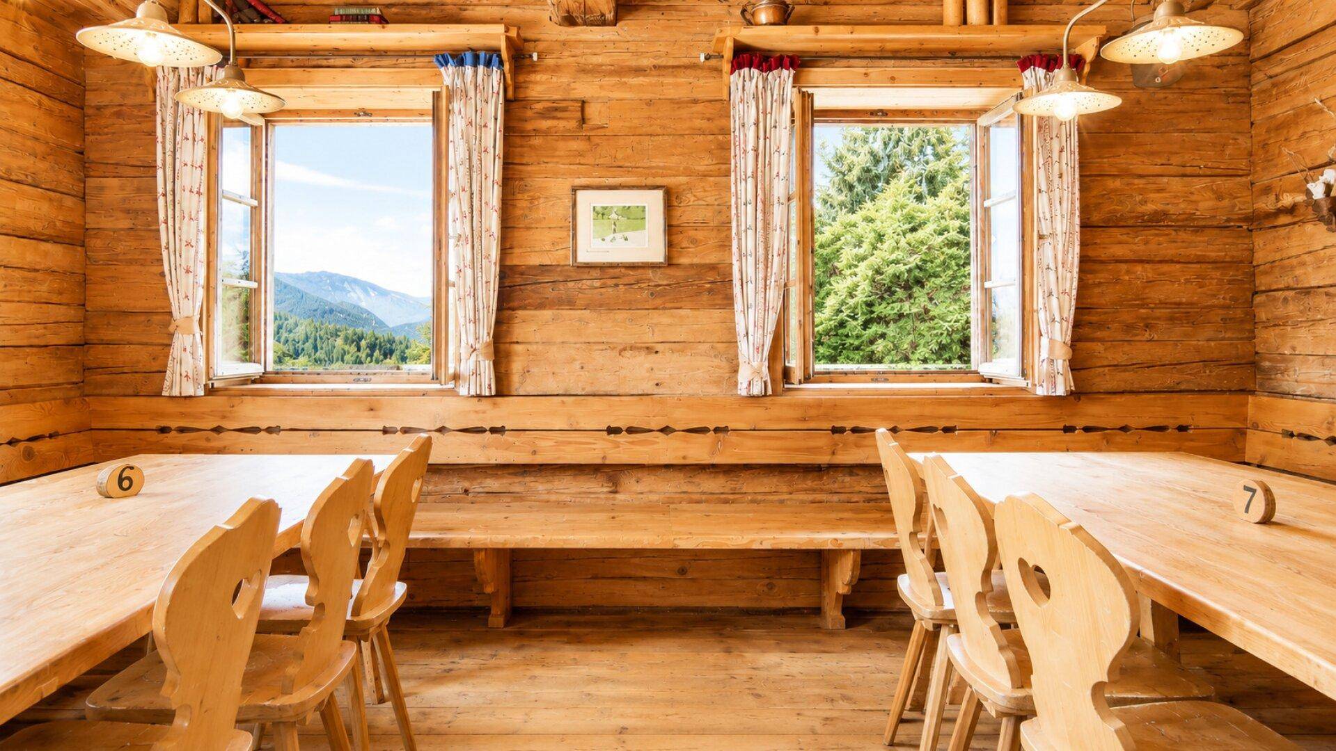 Gemütlicher Essbereich mit Holztischen und Bänken, zwei offenen Fenstern bieten Bergblick und Grün.