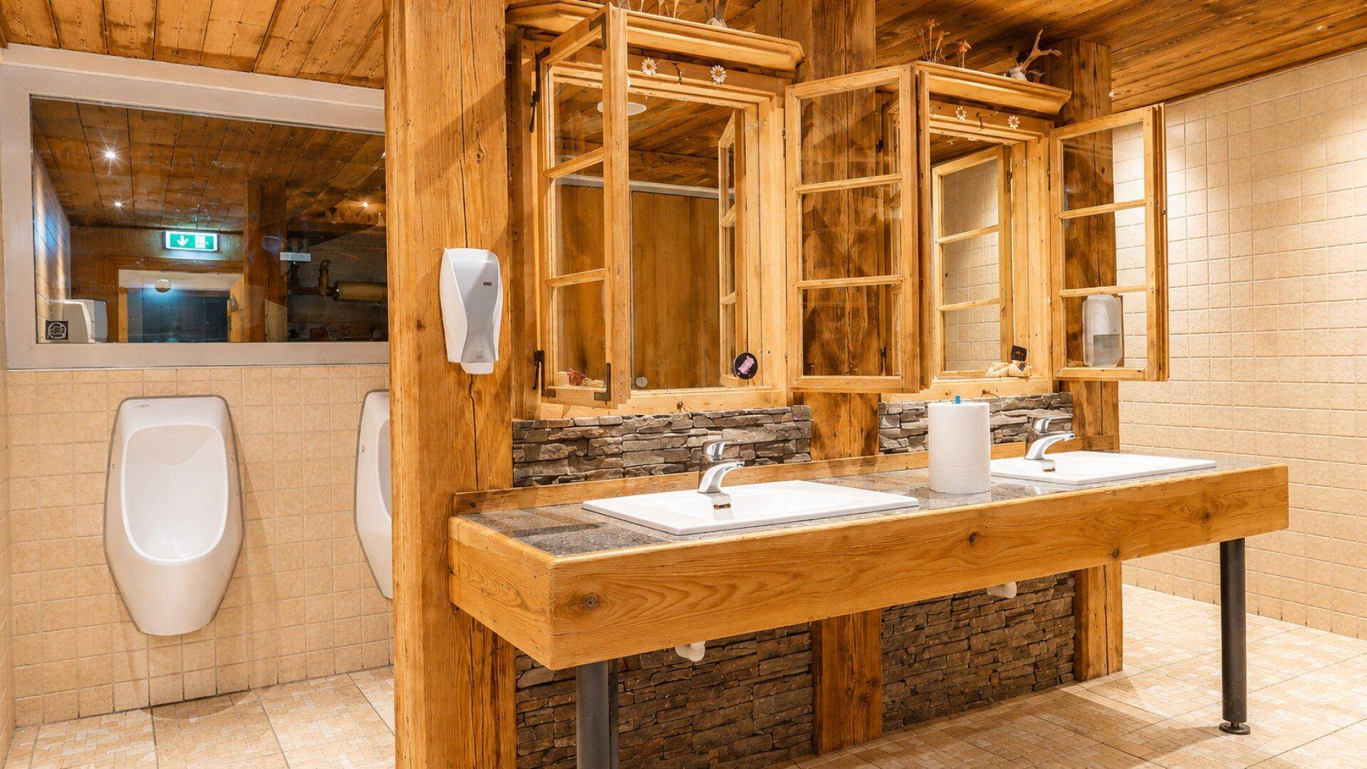Öffentliche Toilette mit zwei Urinalen, Doppelwaschbecken auf Holztheke und rustikalen Holzspiegeln.