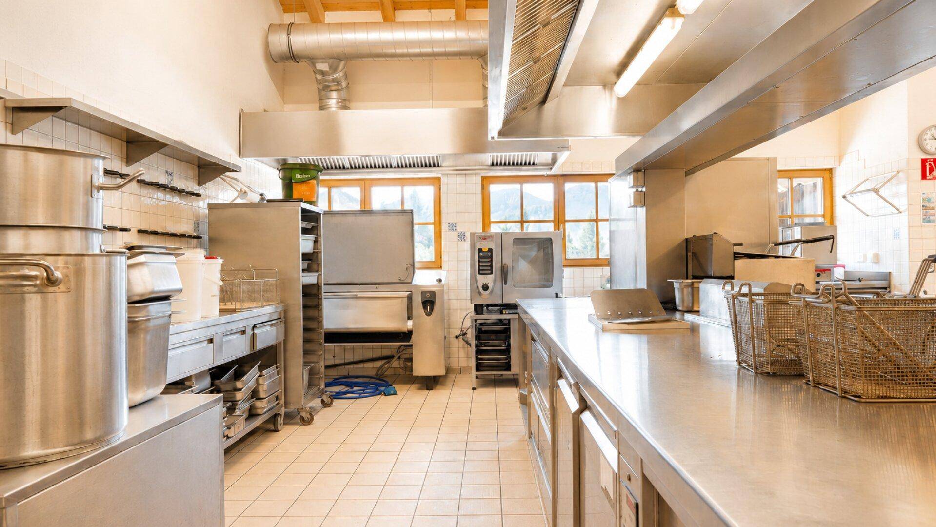 Große Industrieküche mit Edelstahloberflächen, professionellen Geräten und sichtbaren Lüftungsrohren.