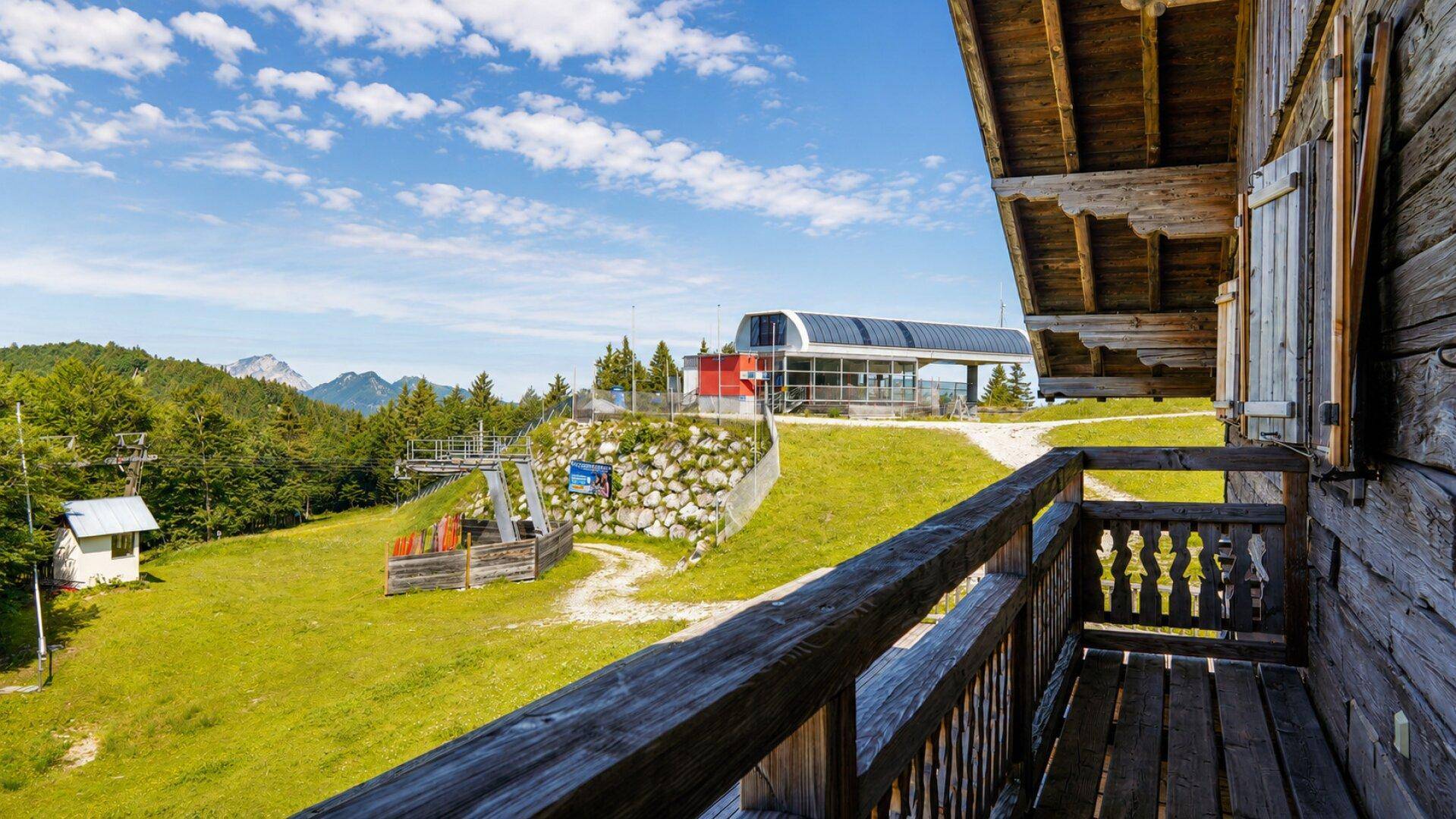 Blick von einem Holzbalkon auf eine grüne Berglandschaft mit einer Seilbahnstation in der Ferne.