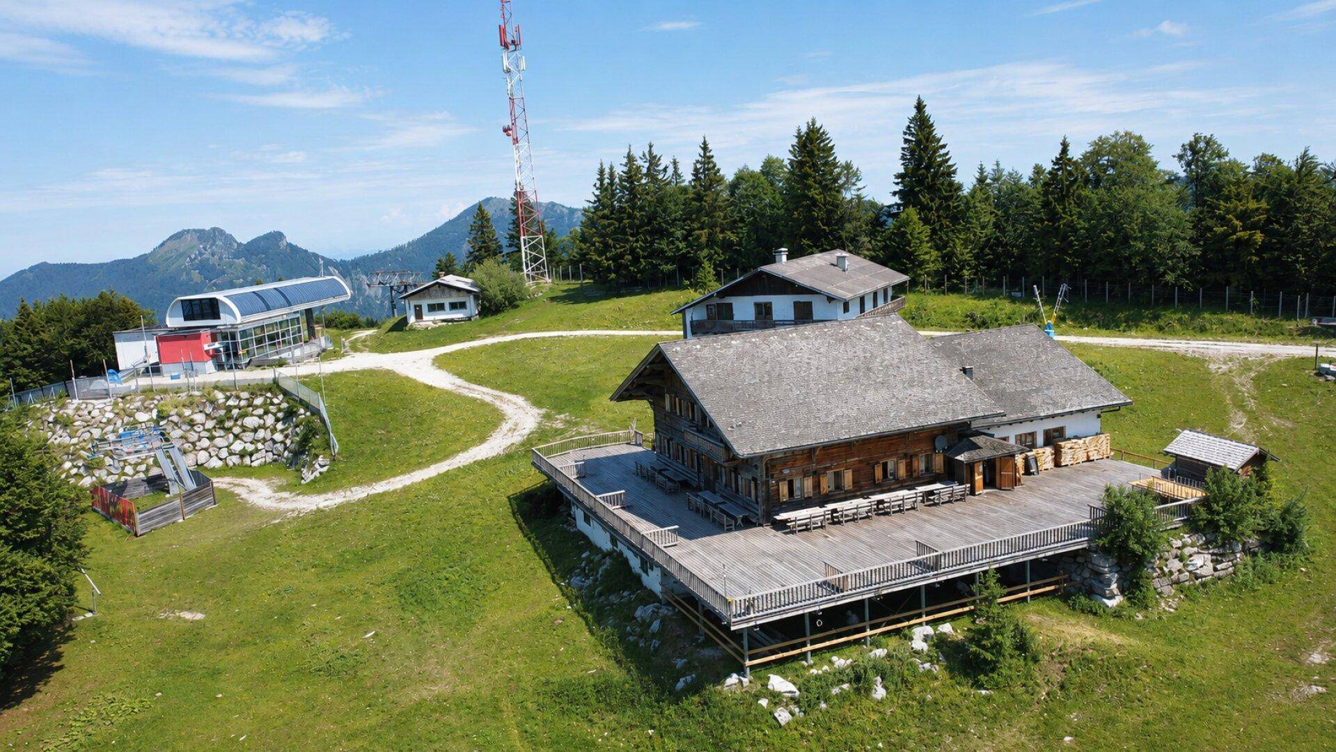 Luftaufnahme des Holzgebäudes mit großer Terrasse, Seilbahnstation und der umliegenden Berglandschaft.