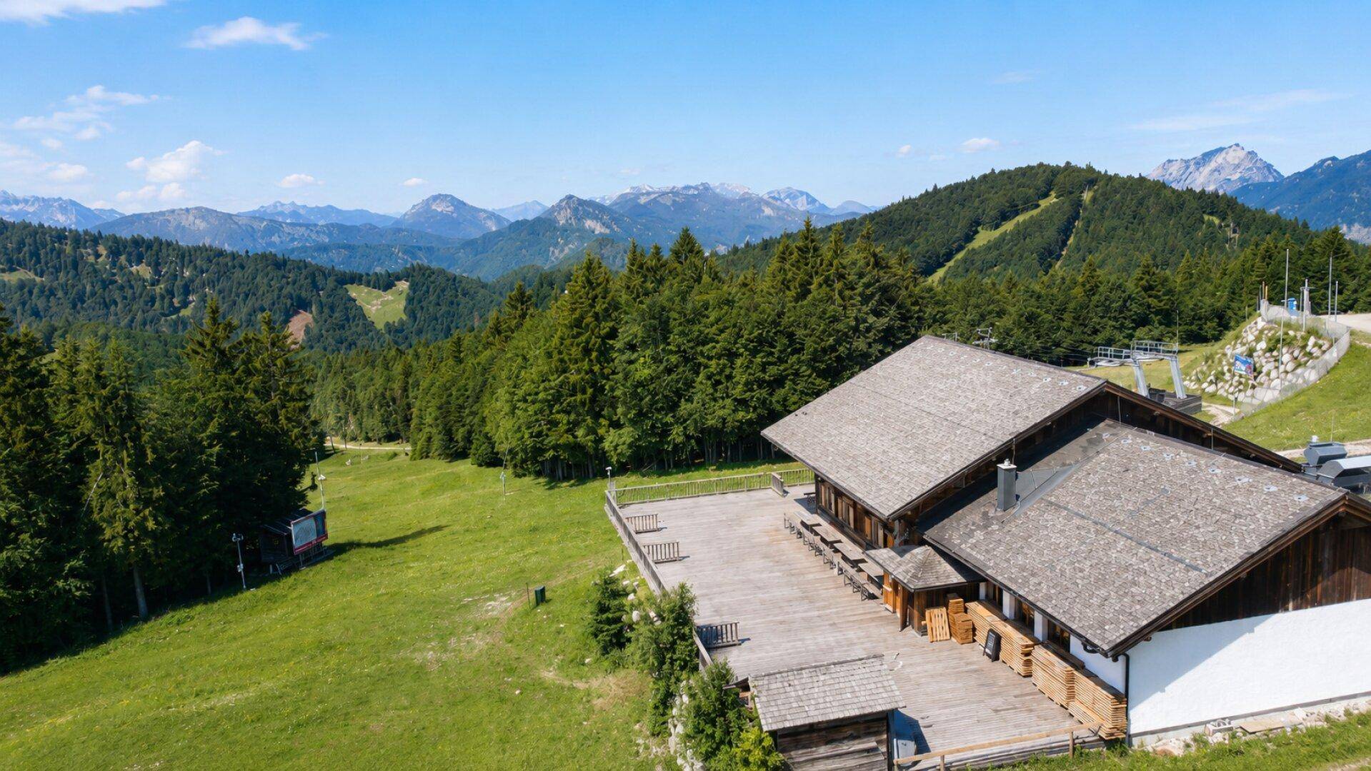 Panoramische Luftaufnahme des Holzgebäudes und der Terrasse, umgeben von weiten Wäldern und Bergen.