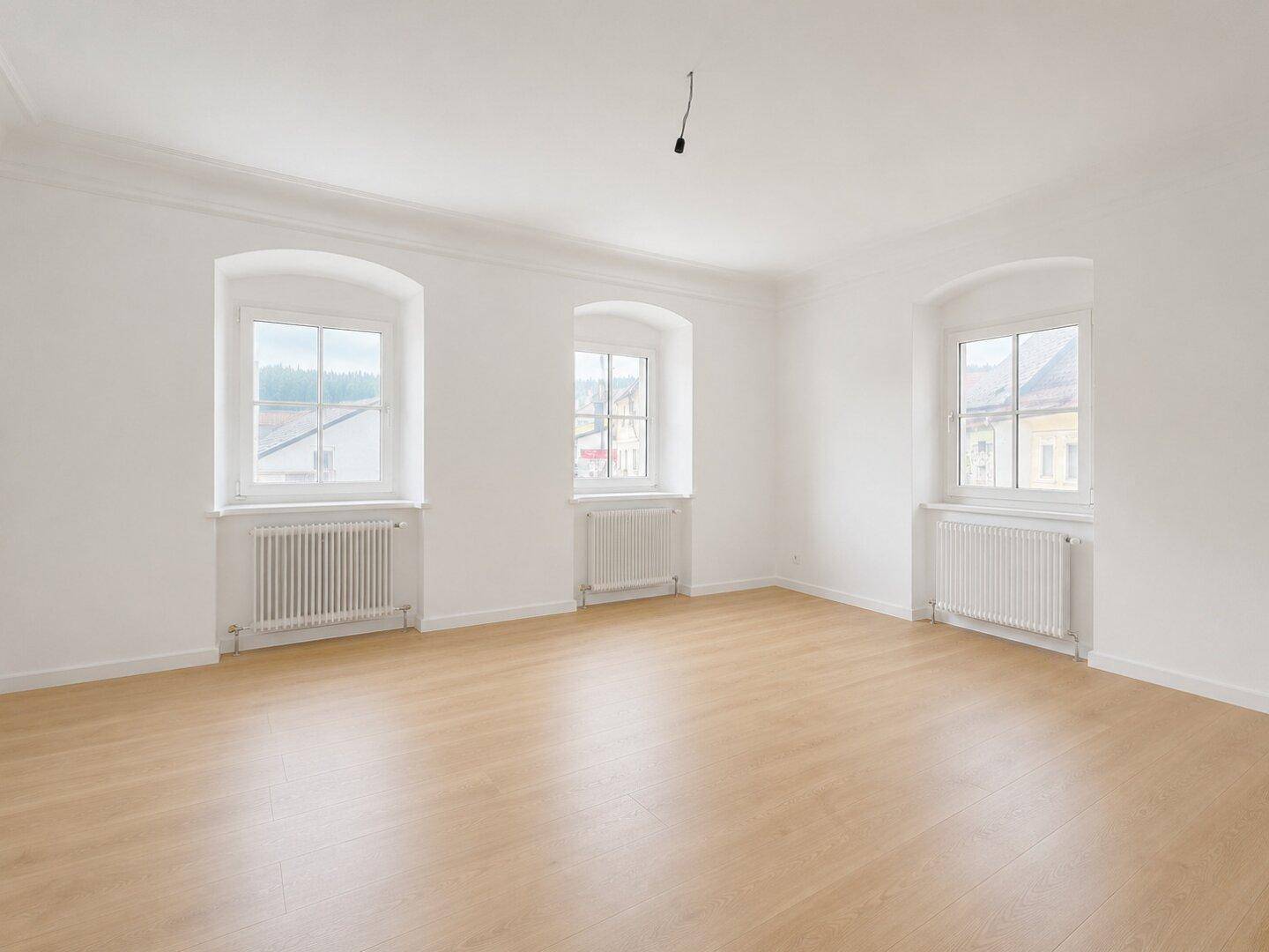 Geräumiges, helles Zimmer mit drei Fenstern, Holzboden und weiß gestrichenen Wänden.