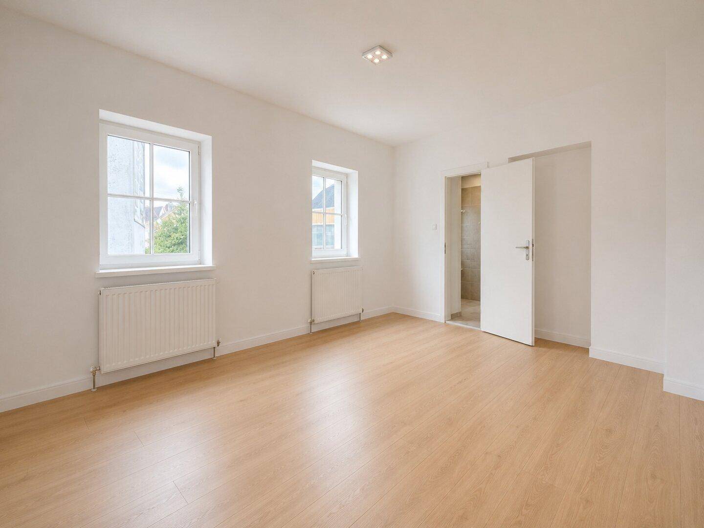 Helles Schlafzimmer mit Holzboden, zwei Fenstern und Zugang zu einem angrenzenden Badezimmer.