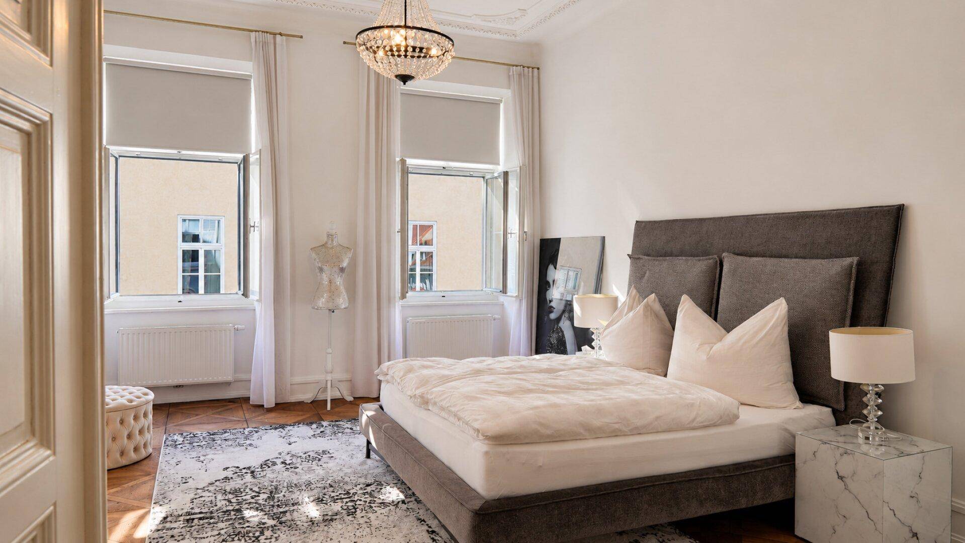 Helles Schlafzimmer mit großem Bett, Parkettboden und zwei hohen Fenstern, die viel Tageslicht hereinlassen.