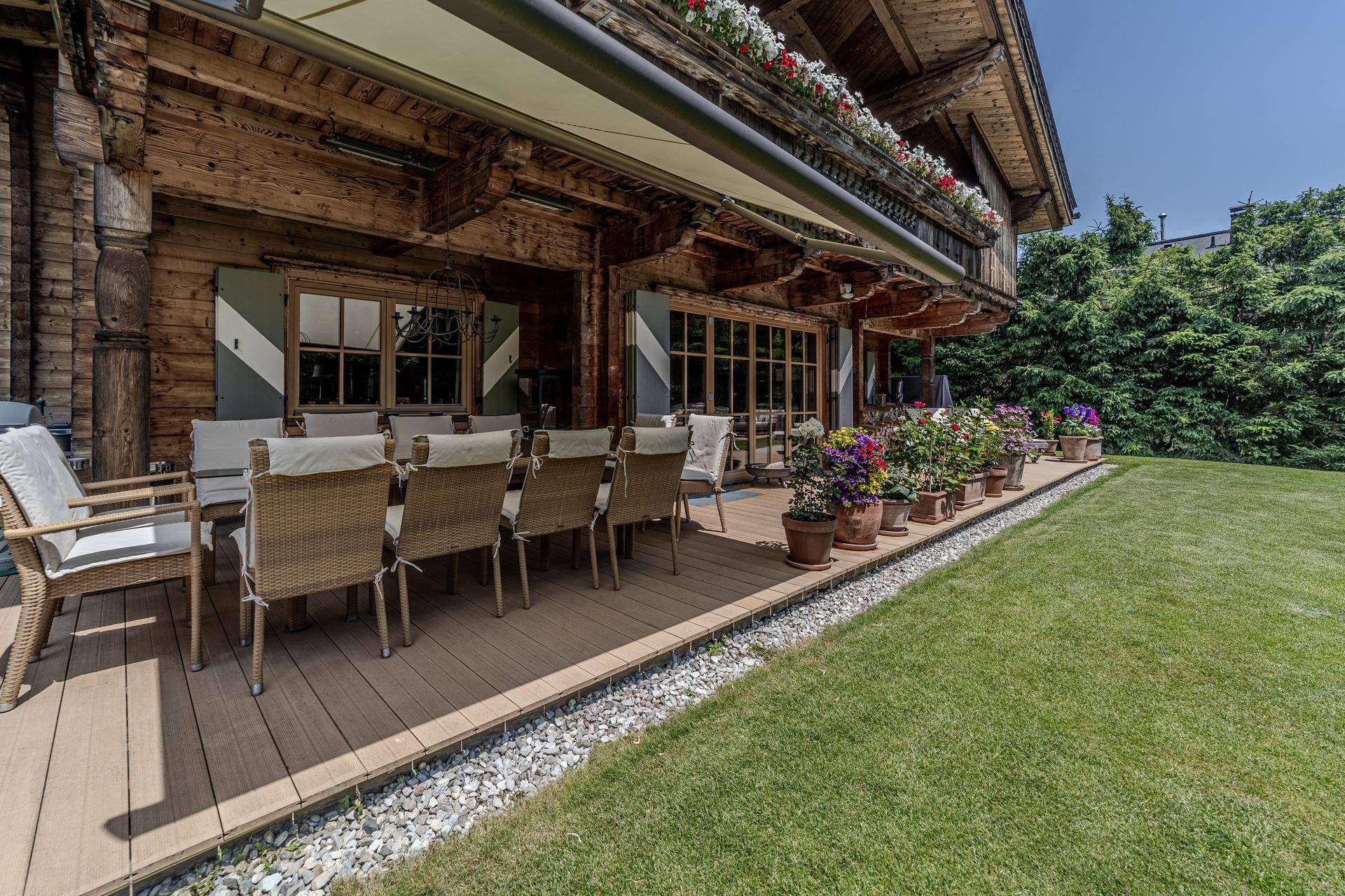 Geräumige Holzterrasse mit großem Esstisch und Stühlen, umgeben von Blumen und Rasenfläche.