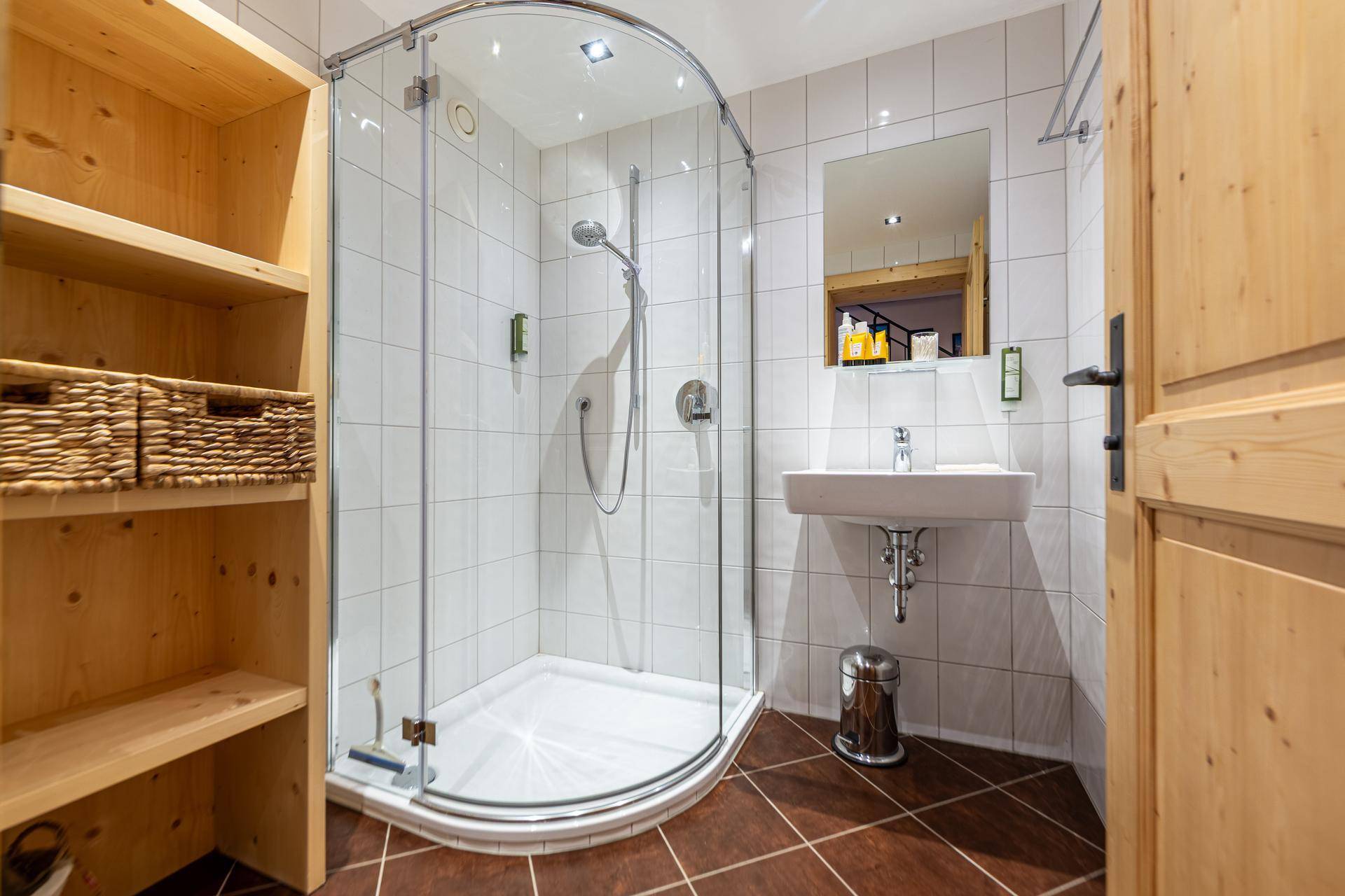 Kleines Badezimmer mit Dusche, Waschbecken und weißen Fliesen, sowie Holzregalen.