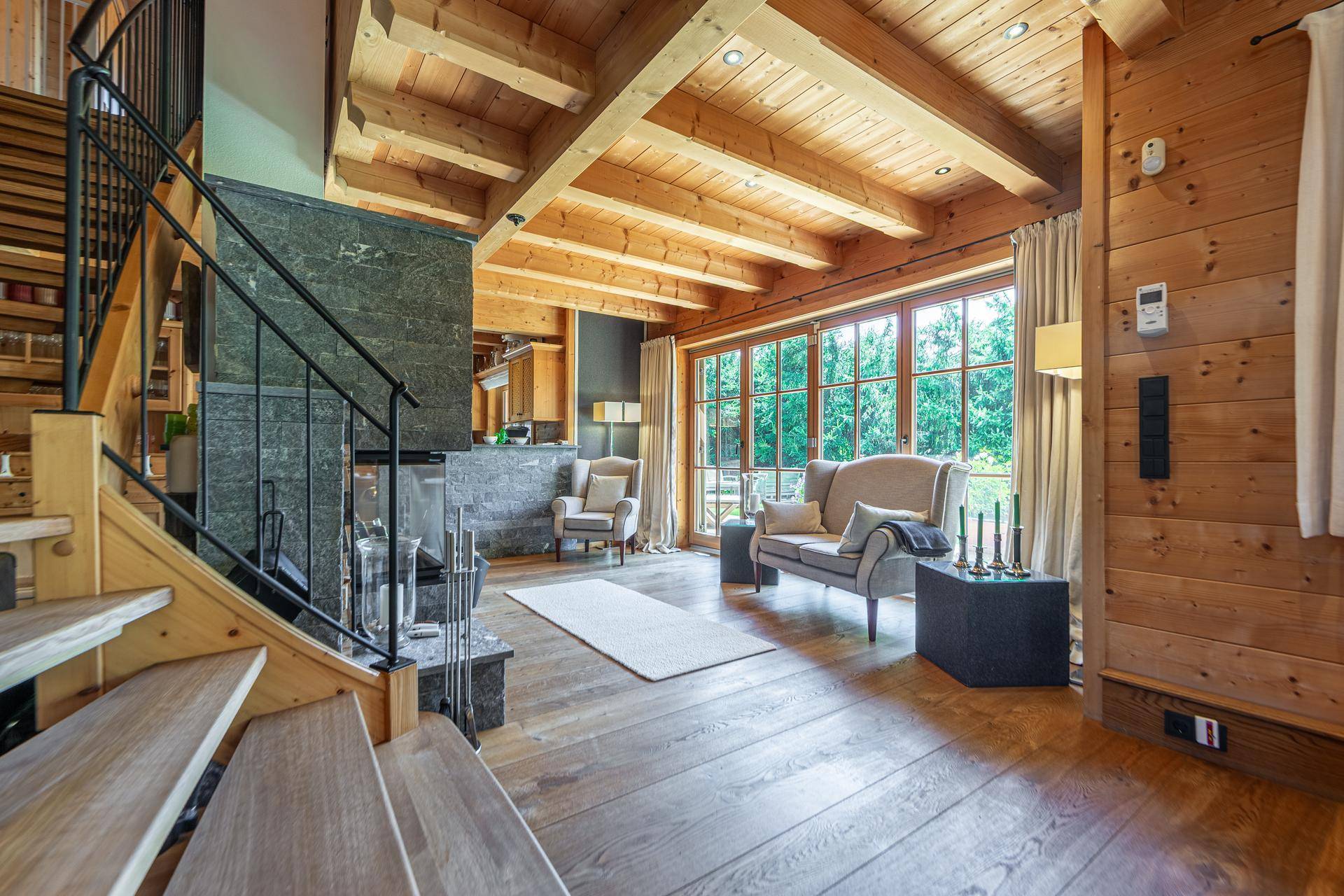 Wohnzimmer mit Kamin, Holztreppe zur Galerie und großen Fenstern mit Blick ins Grüne.